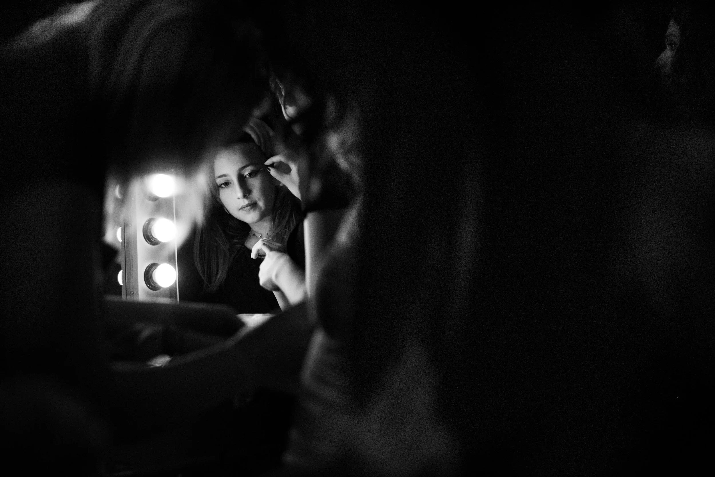 A woman looking at her reflection in a mirror, touching up her makeup with vanity lights around the mirror illuminating her face, in a black and white photograph.