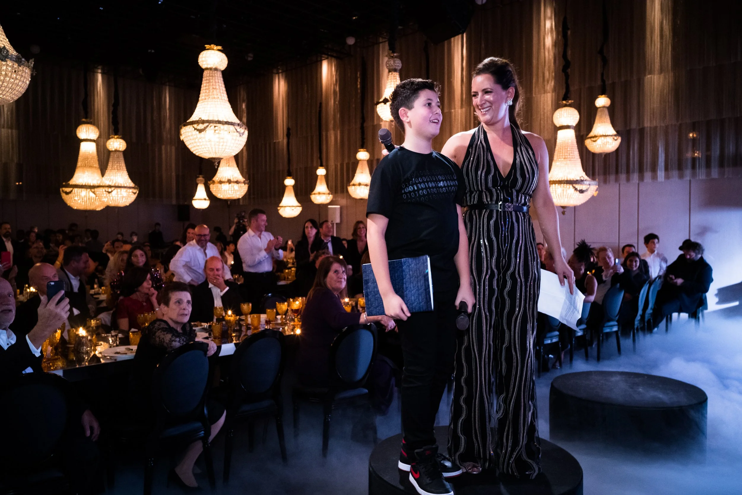 A woman and a young boy stand on a small stage at a formal event, smiling at each other. The woman is dressed in a striped jumpsuit, and the boy is wearing a black t-shirt and pants. The boy holds a folder, and the woman holds a paper, with a microph