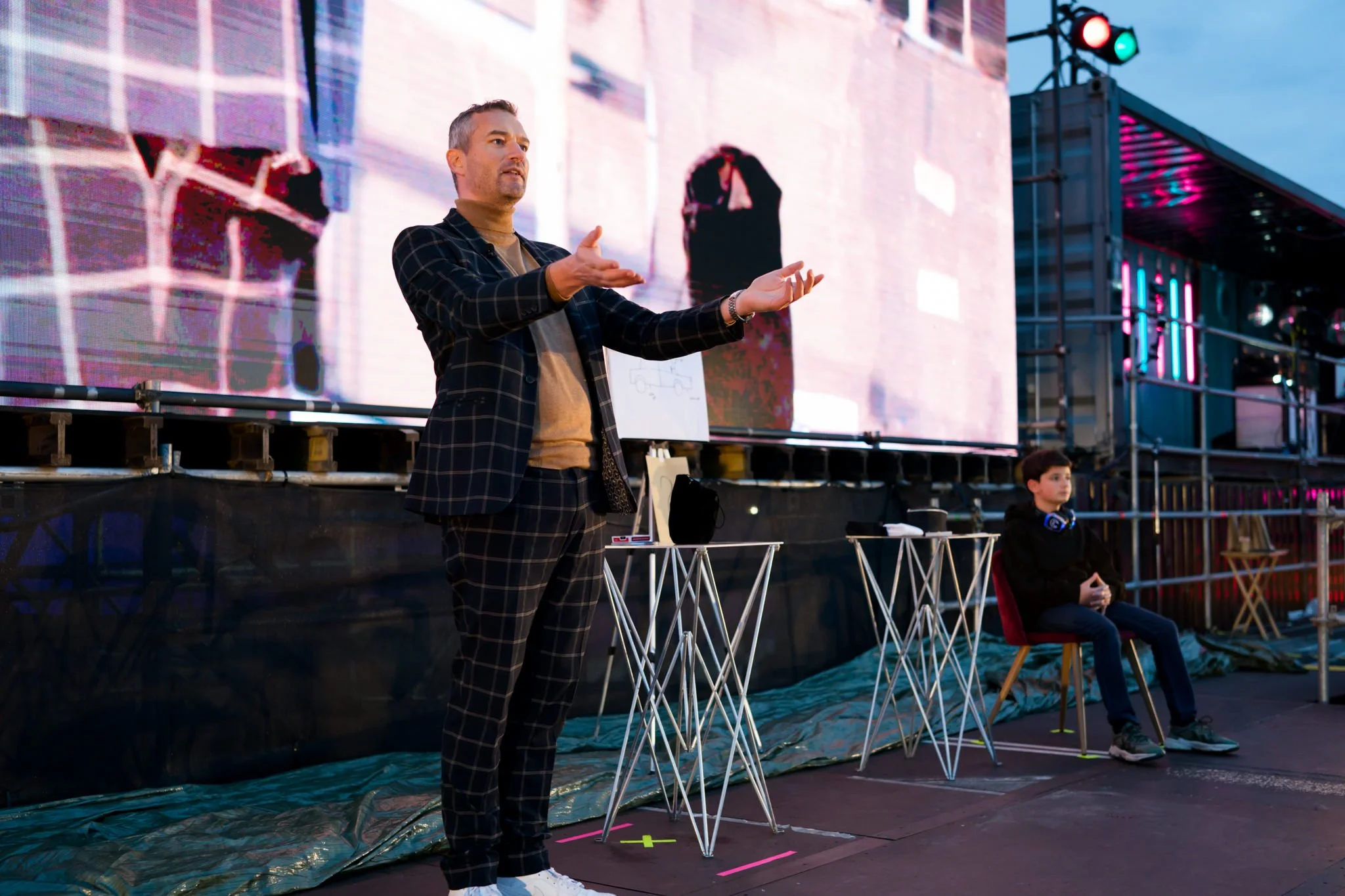 A man in a checkered suit giving a presentation in front of a large screen, with a young boy sitting nearby. The scene appears to be outdoors with a stage and neon lights.