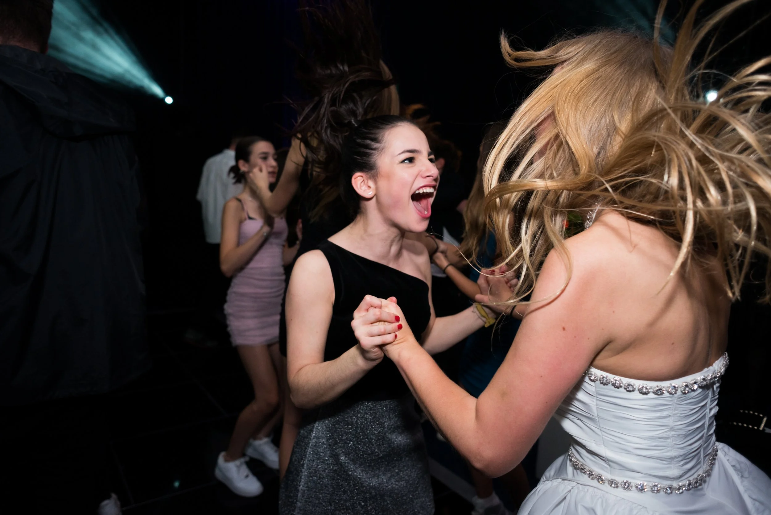 Two women dancing and holding hands at a party or celebration, surrounded by other people on a dance floor.