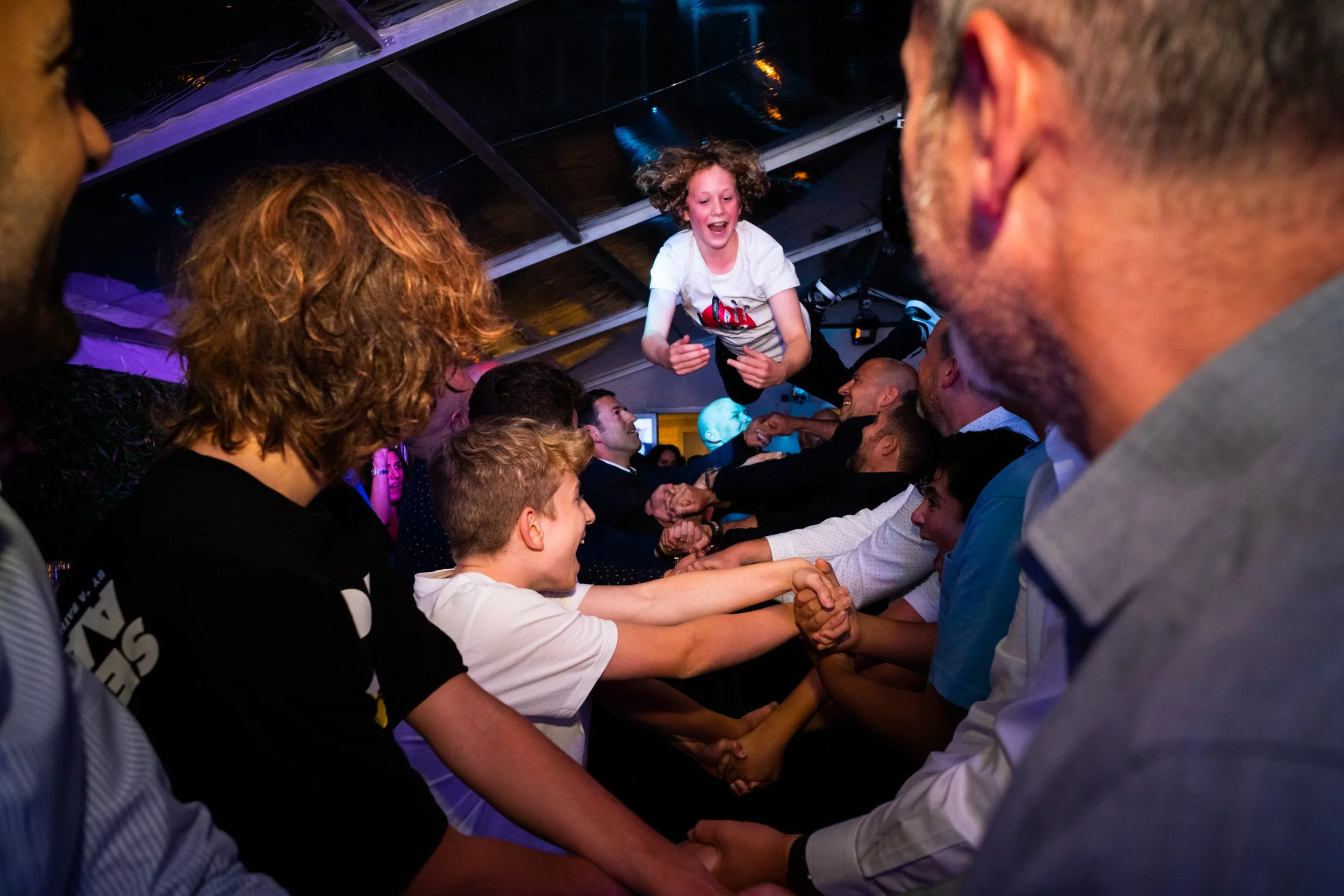 A group of people holding hands in a circle under a tent, with a girl jumping or being lifted in the center during a celebration or event.