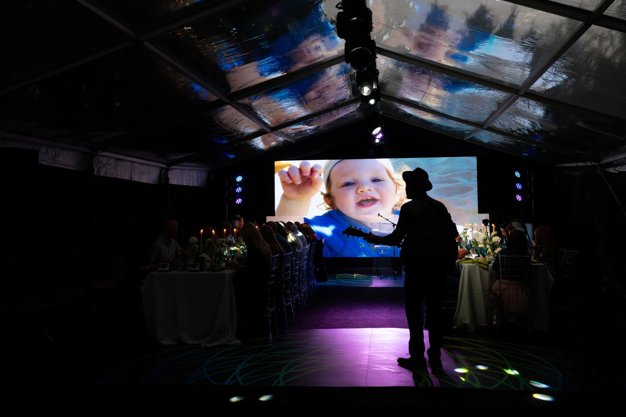 A person playing guitar at a reception with a large screen showing a close-up photo of a smiling child waving.