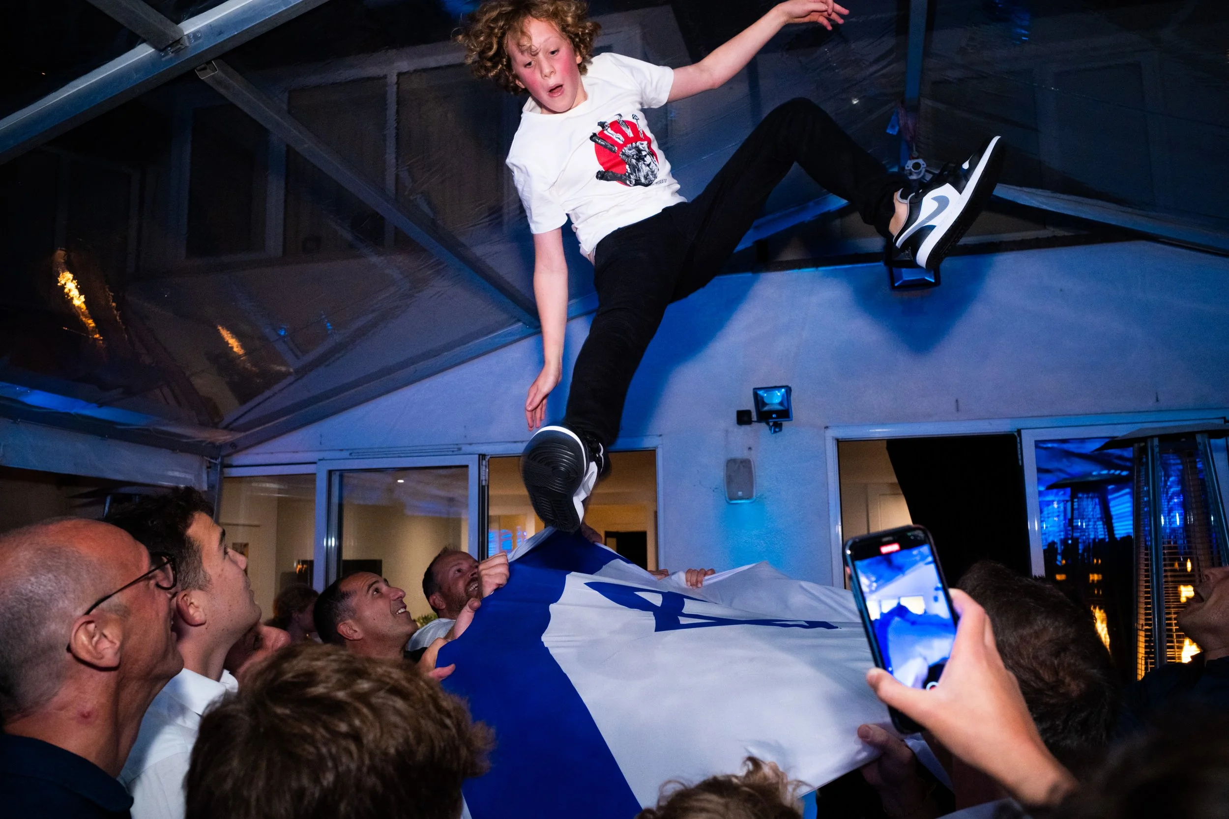 A young boy is being lifted into the air at a celebration or party, surrounded by adults, with one person holding a flag. The scene is lively and festive.