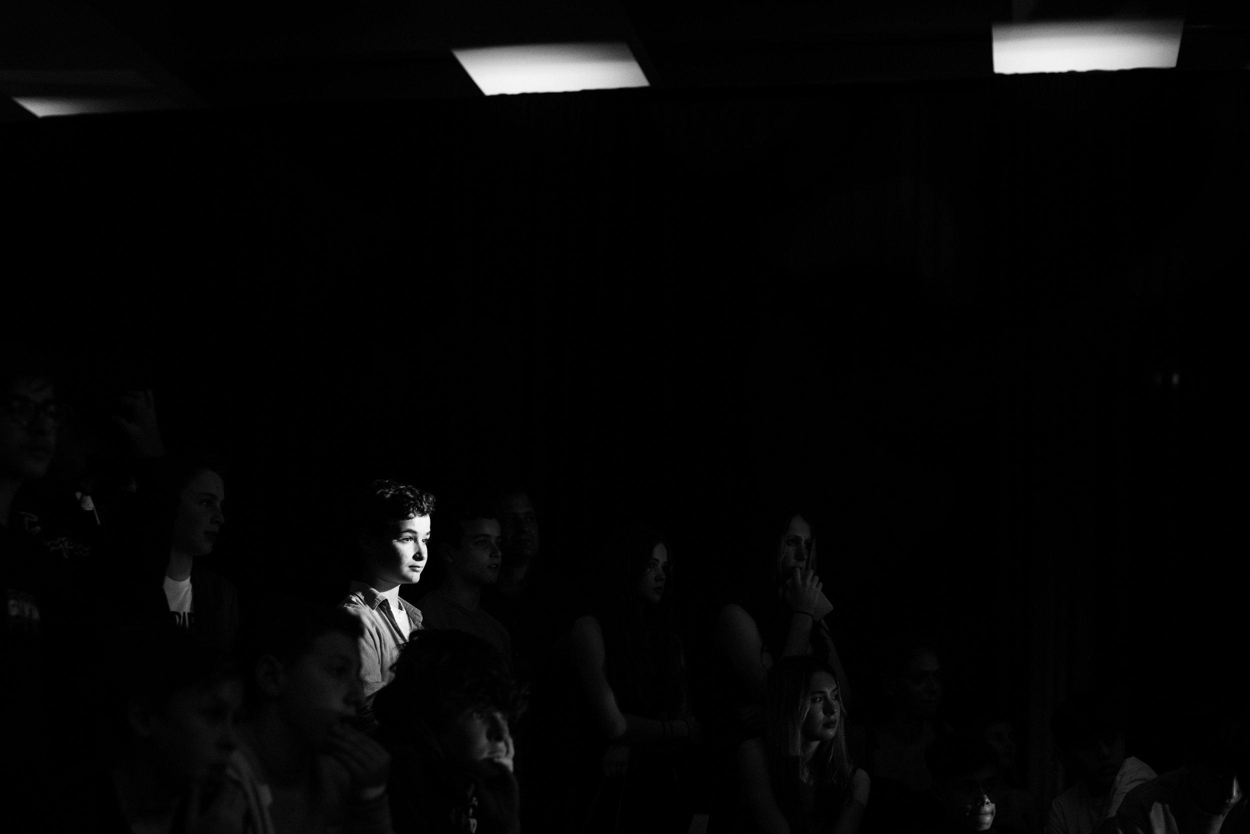 Audience of people watching a performance, with one young woman illuminated by a spotlight.