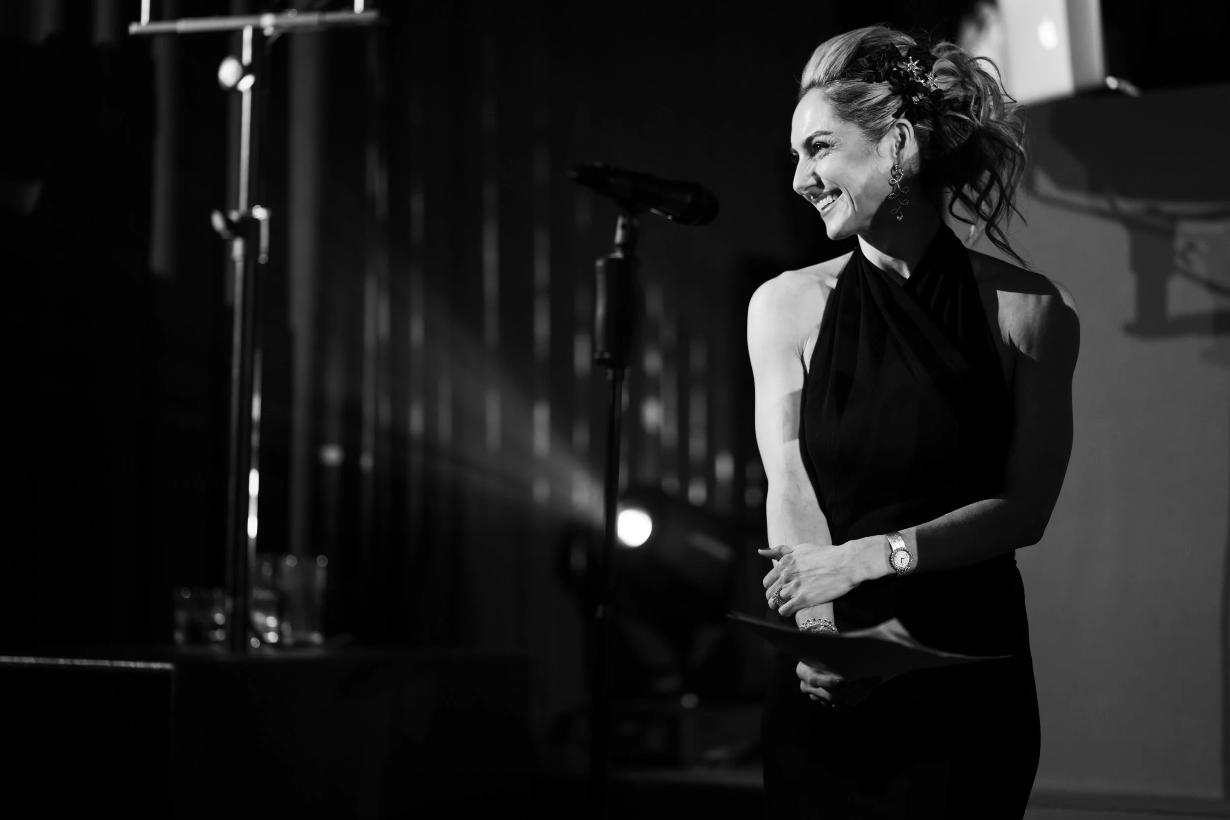 A woman in a black dress standing next to a microphone on stage, smiling and looking to her right, with her hands clasped in front of her, in a black and white photograph.