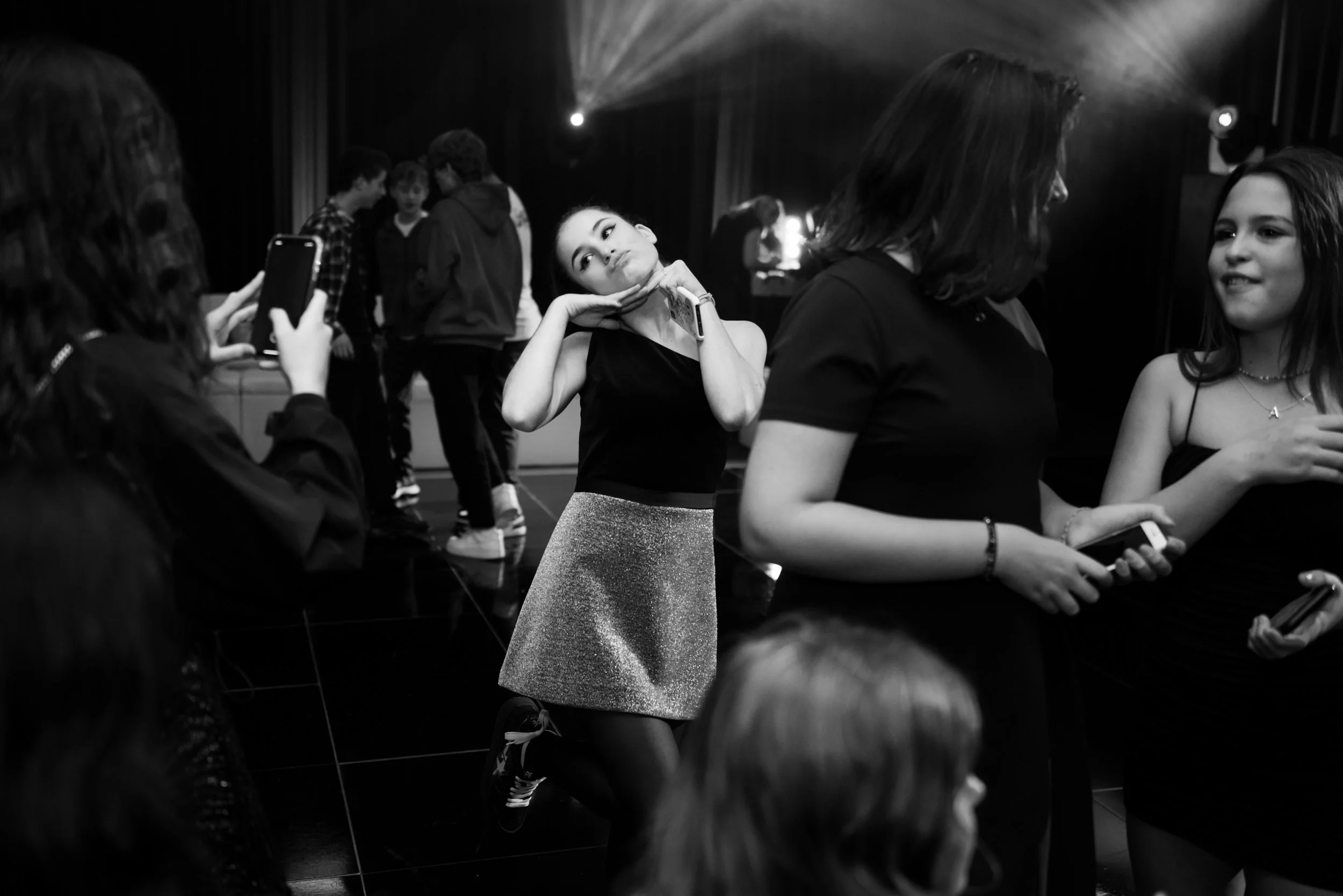 Young girl making a playful face while posing with her hands under her chin at a group gathering or party with other girls and teenagers.