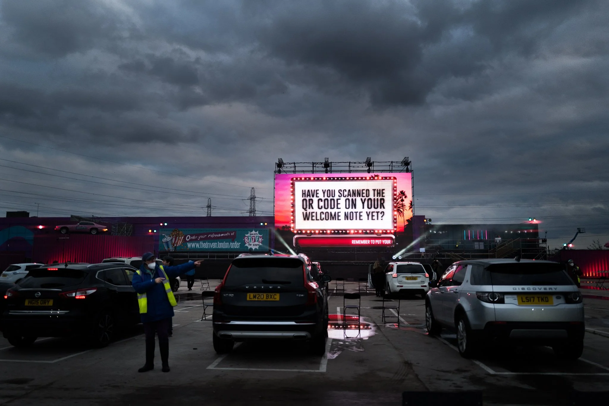 A drive-in theater parking lot during dusk with a large illuminated screen displaying the message, 'Have you scanned the QR code on your welcome note yet?' Multiple cars are parked, and a person wearing a face mask and a reflective vest is standing i