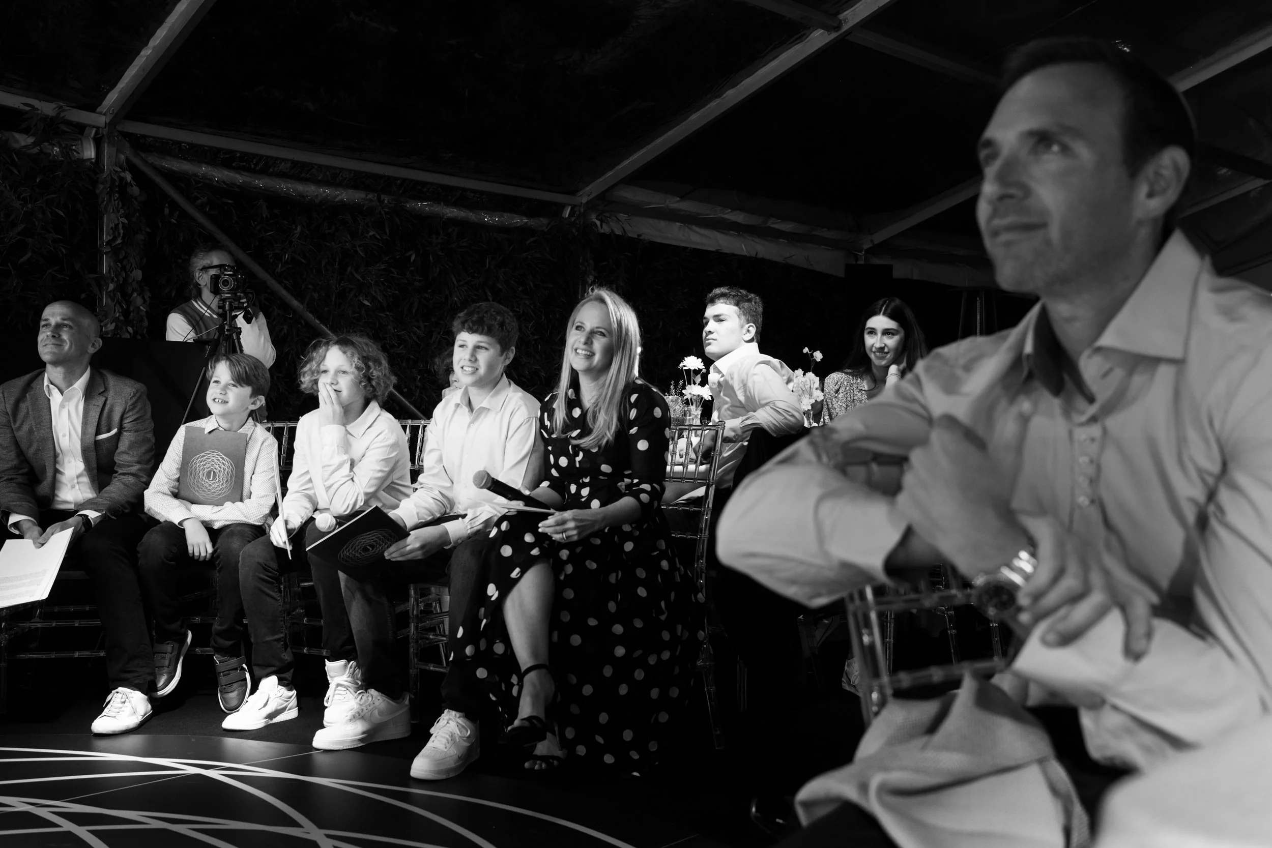 People seated at an event, some holding notebooks, with a photographer in the background, in a tent or covered outdoor area.