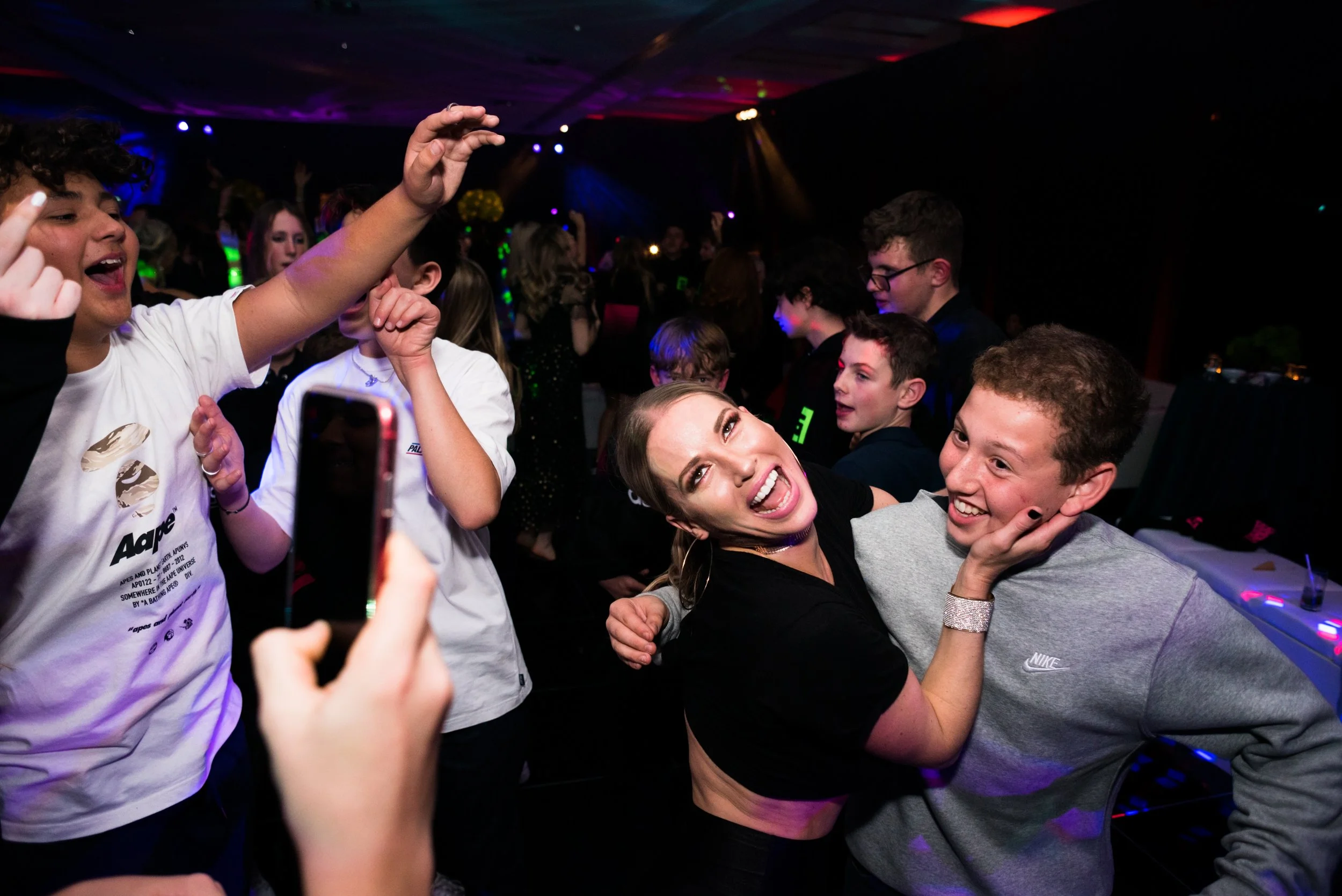 A lively party scene with a group of young people dancing and taking pictures. In the foreground, a woman is hugging a young man, both smiling and enjoying the moment. The background shows more people dancing under colorful party lights.