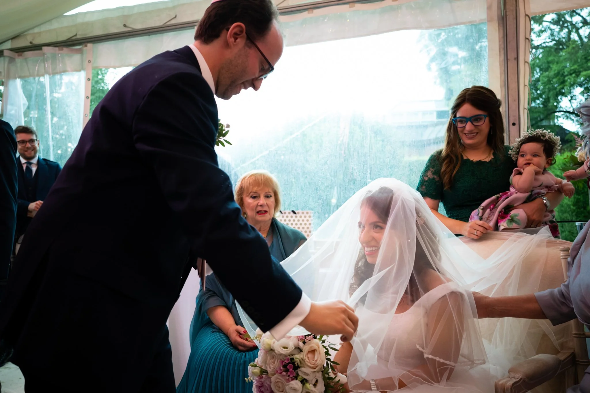 A bride with a veil smiling as a man adjusts her veil at a wedding ceremony inside a tent, with women and a baby girl in the background.