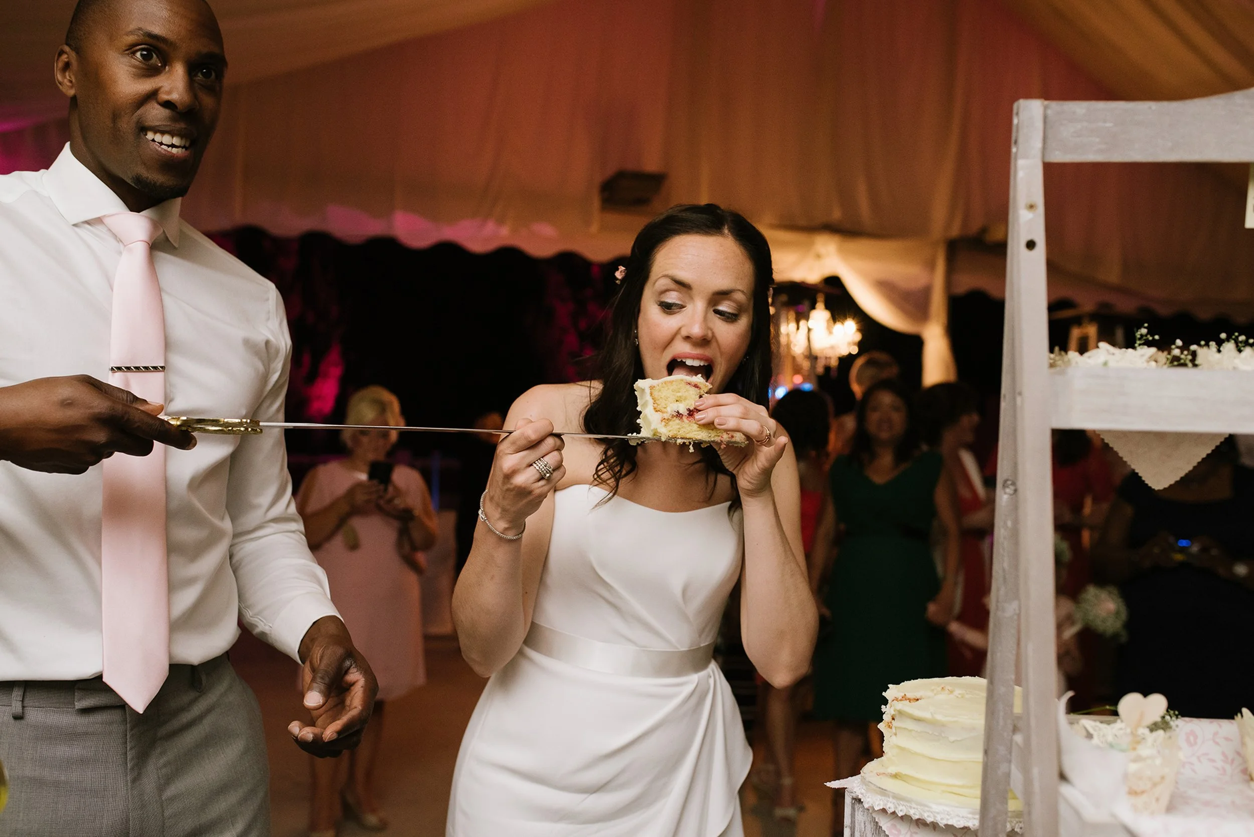 Woman in a white dress cutting a piece of cake with a sword at a wedding reception, standing next to a man in a white shirt and pink tie, with guests in the background.