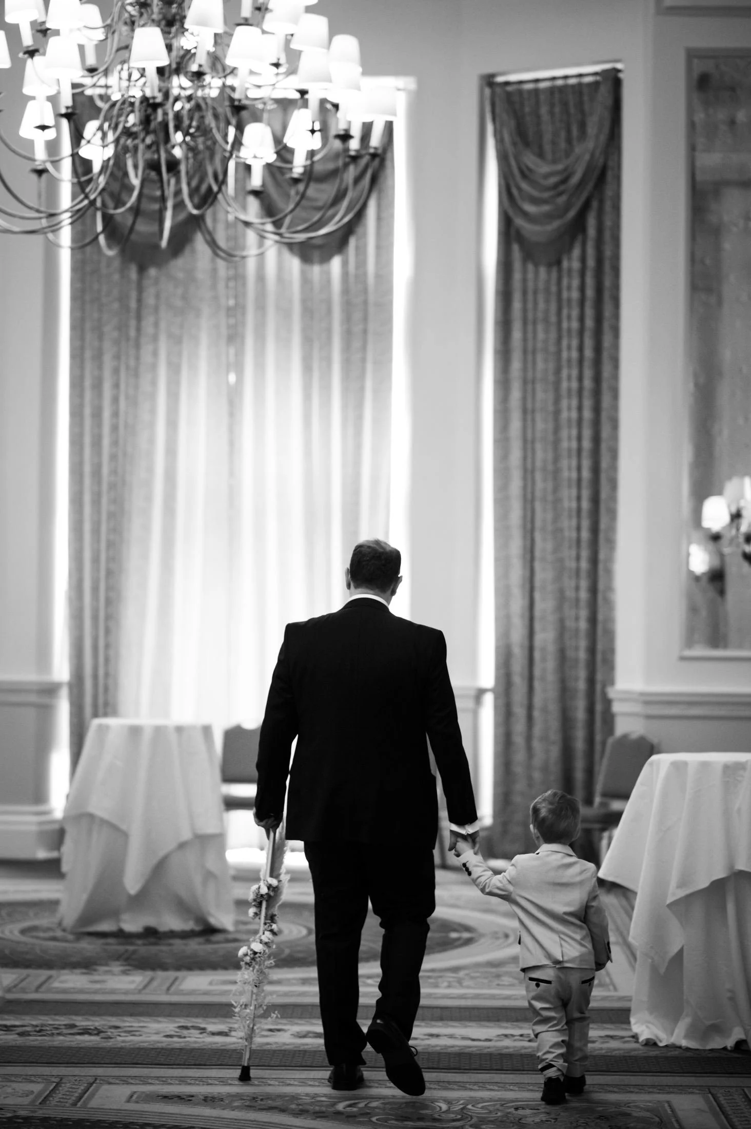 A man in a suit walking with a young boy in a fancy room, holding hands, with elaborate drapes, tables, and a chandelier, all in black and white