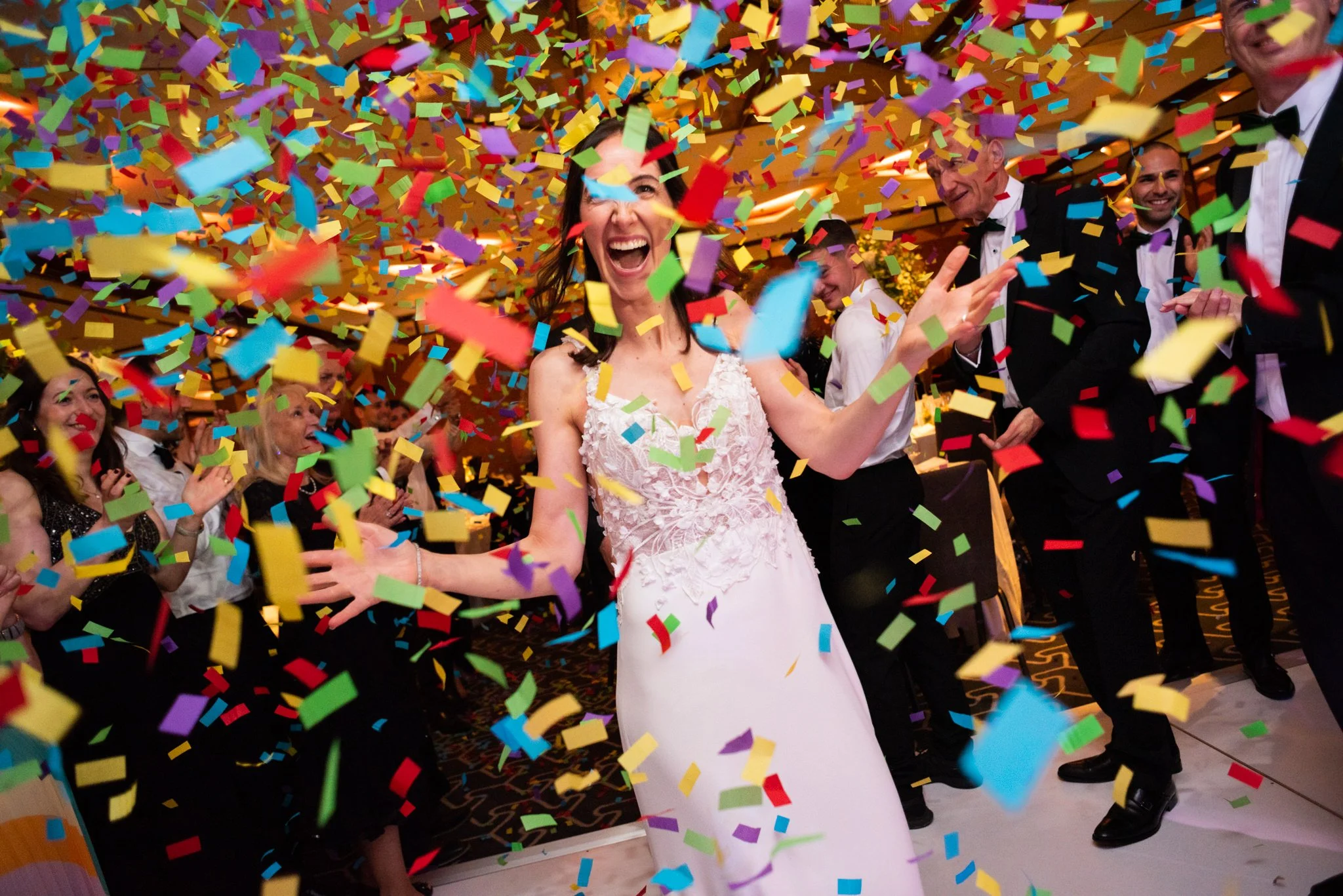 A woman in a white wedding dress celebrating with colorful confetti falling around her at a wedding reception, surrounded by smiling guests in formal attire.