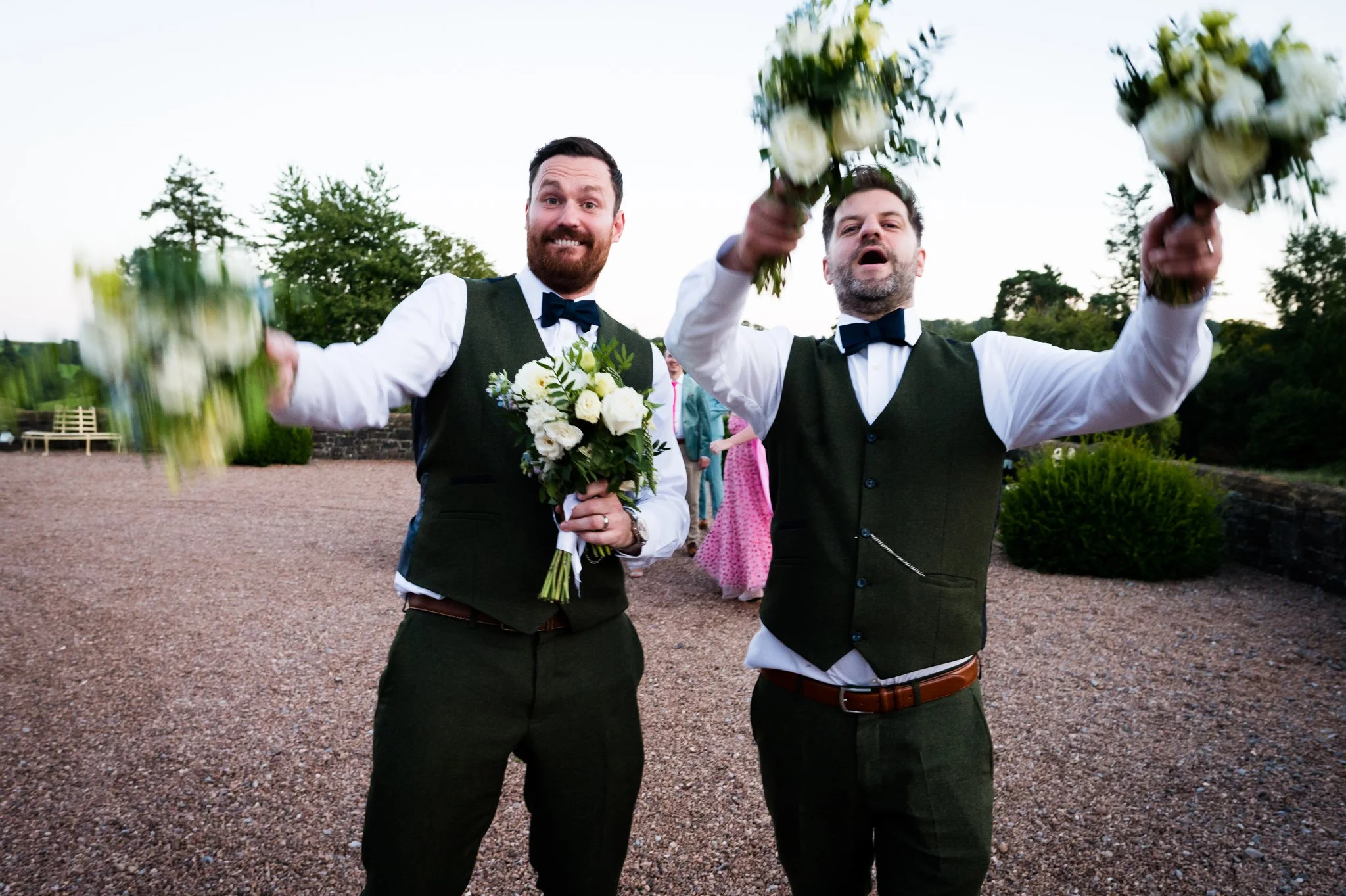 Two men in formal wedding attire holding bouquets of flowers, smiling, and celebrating outdoors.