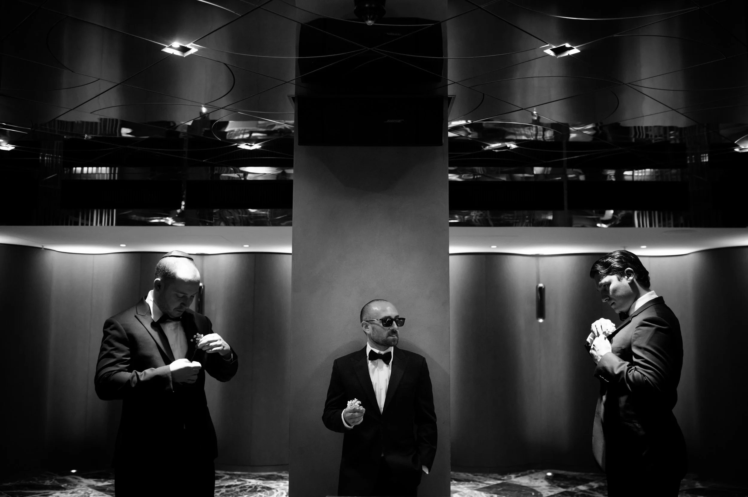 Three men in tuxedos stand in an elegant, dimly lit interior with reflective ceiling panels. The man on the left is looking down, holding a boutonniere, the man in the center, wearing sunglasses, is looking to his right, also holding a boutonniere, a