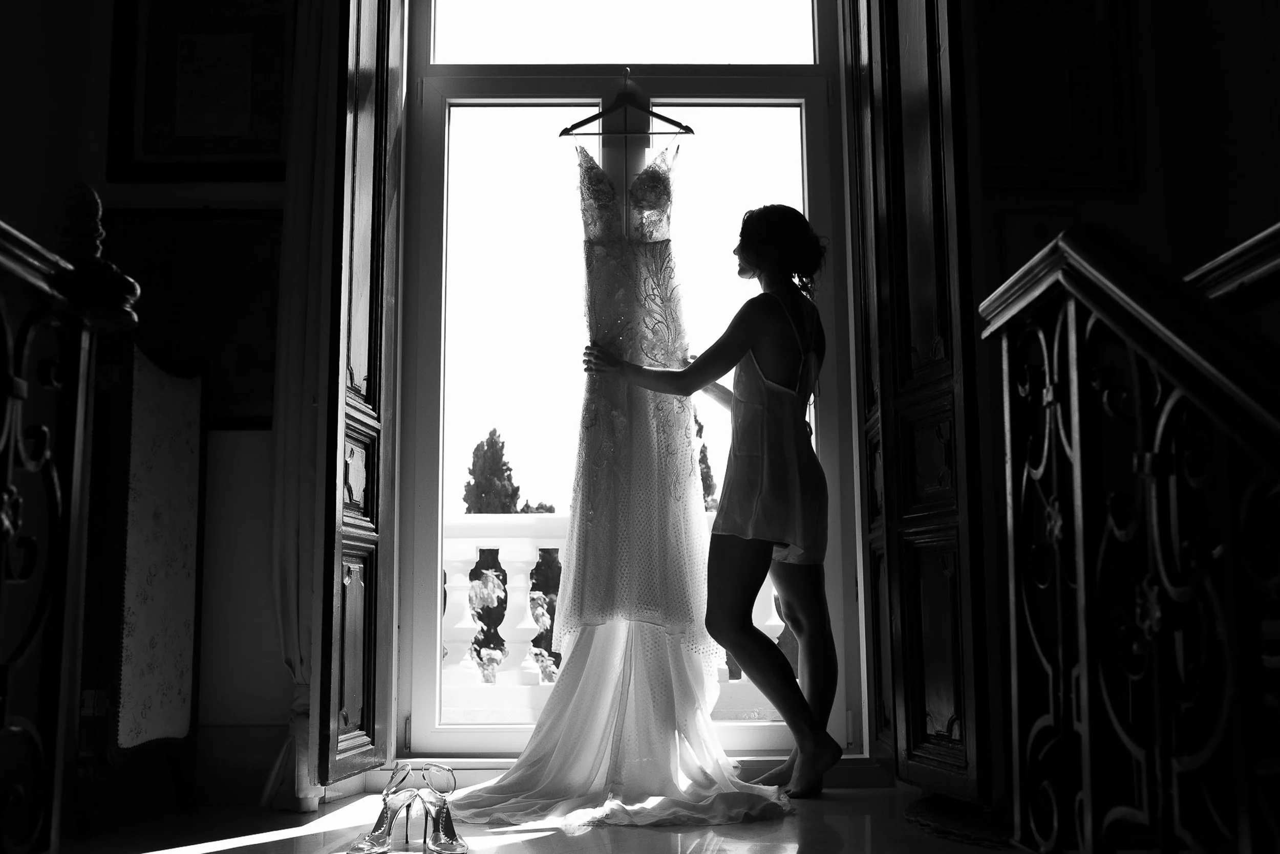 A woman looking at a wedding dress hanging by a window. The photo is in black and white, with natural light coming through the window.