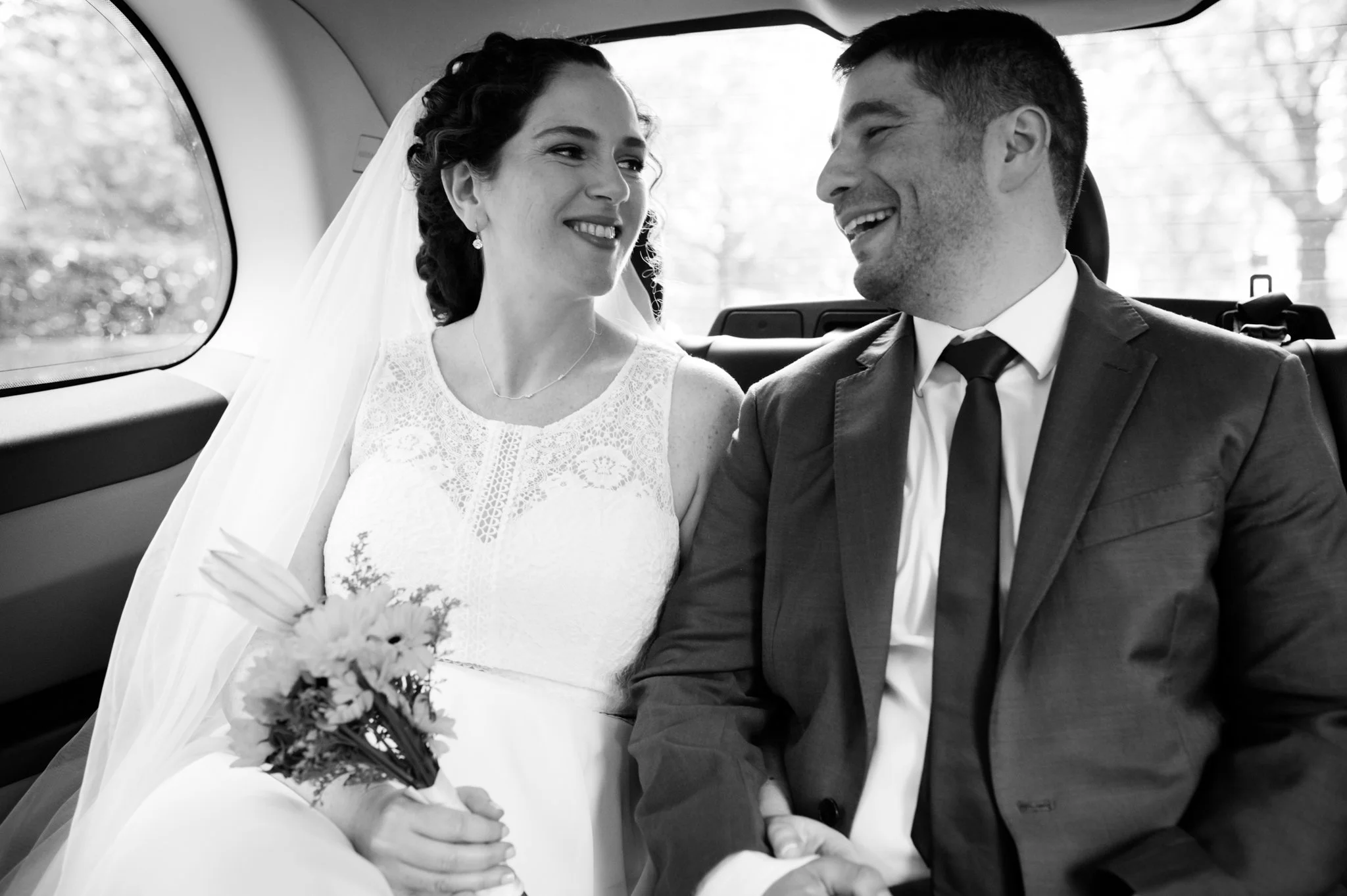 A black and white photo of a bride and groom sitting in a vehicle, smiling and looking at each other. The bride is wearing a lace wedding dress and holding a small bouquet. The groom is dressed in a suit and tie.