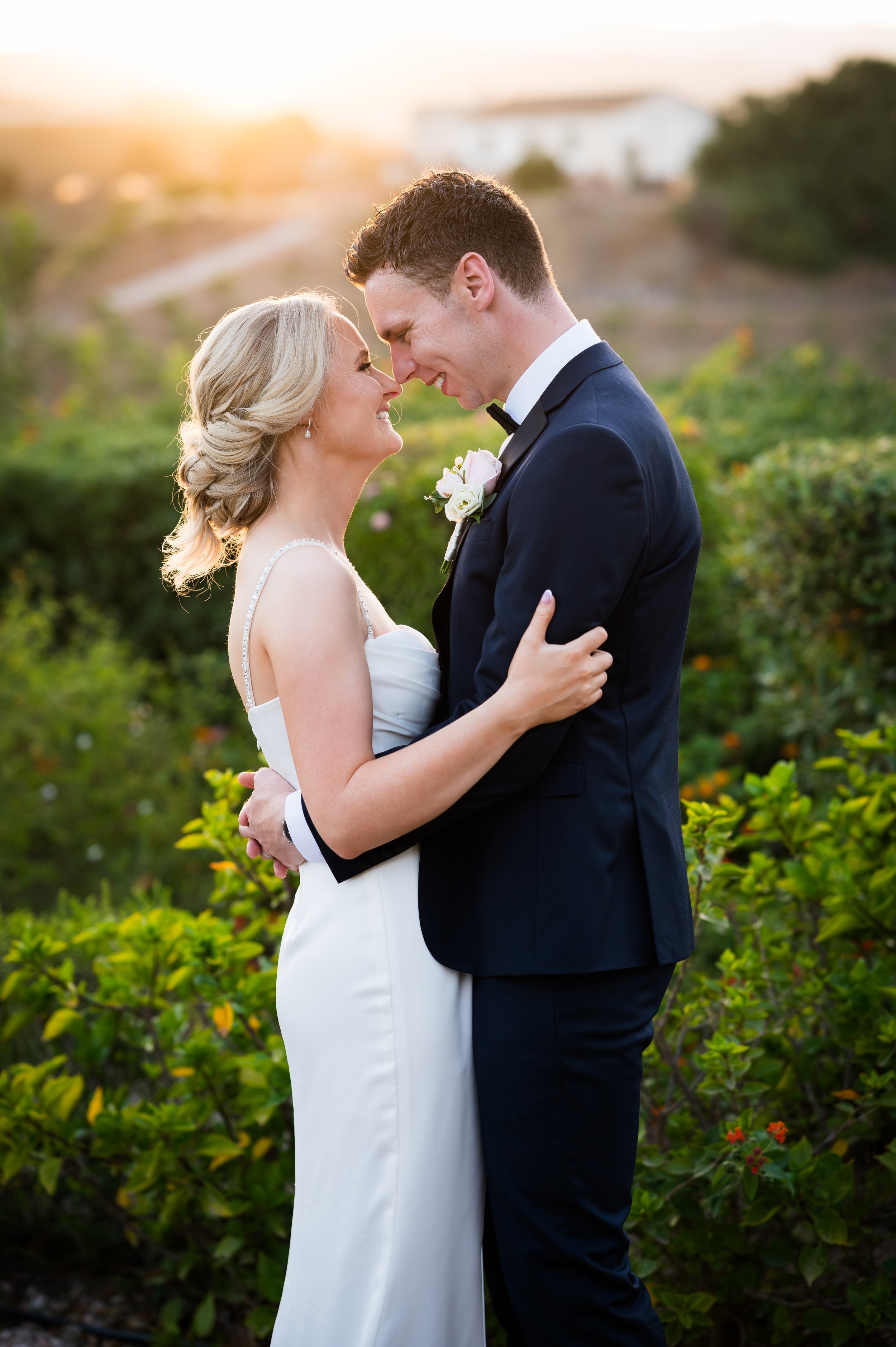 A bride and groom embrace outdoors at sunset, smiling and touching foreheads, with greenery and a blurred background.