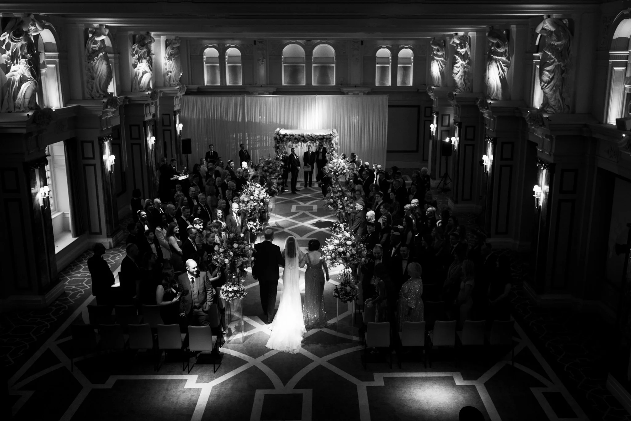 A black and white photo of a wedding ceremony in a grand hall, with the bride and groom walking down the aisle surrounded by guests and floral arrangements.