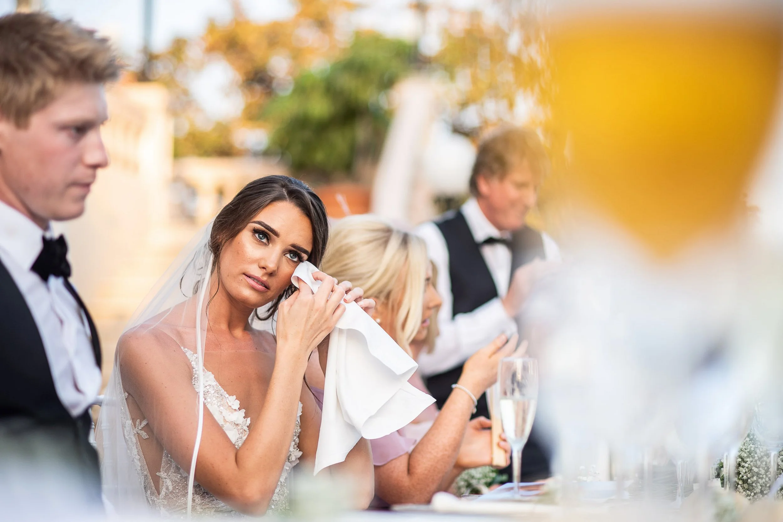 A bride wiping tears with a tissue at a wedding reception with guests sitting at the table, including a man and a woman, and an outdoor setting with trees and warm lighting.