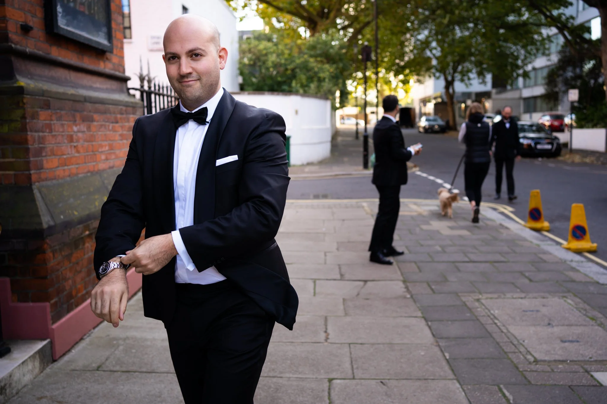 A man in a black tuxedo and bow tie is adjusting his watch on a city sidewalk, with other people in formal attire and a dog in the background.