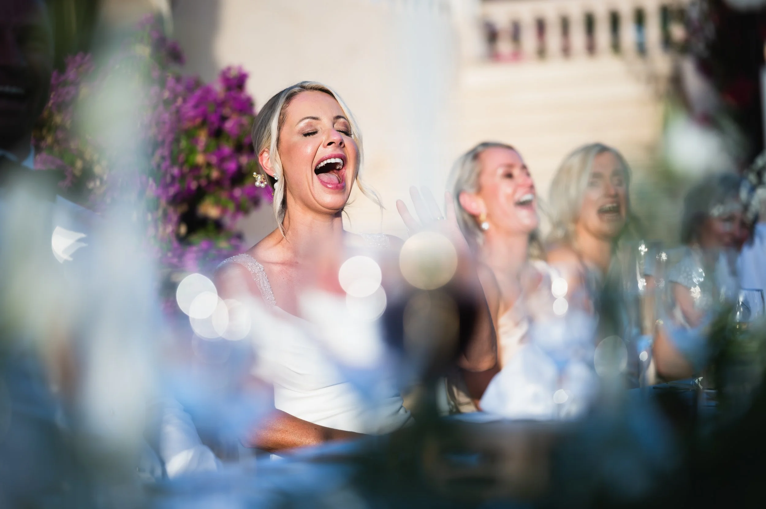 Group of women sitting at a table, laughing and enjoying a celebration outdoors with flowers in the background.