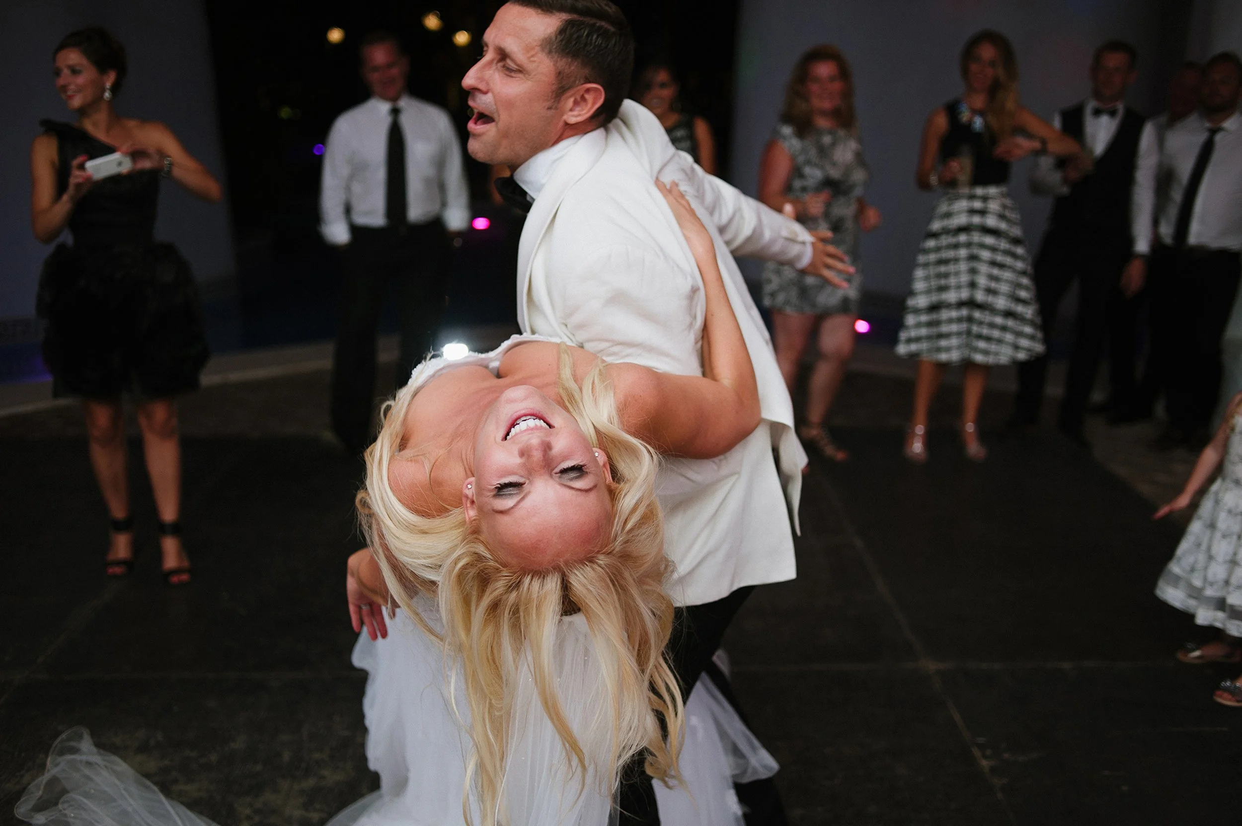 A newlywed couple sharing a dance at their wedding reception, with the groom in a white tuxedo and the bride in a white wedding dress, surrounded by guests in formal attire.