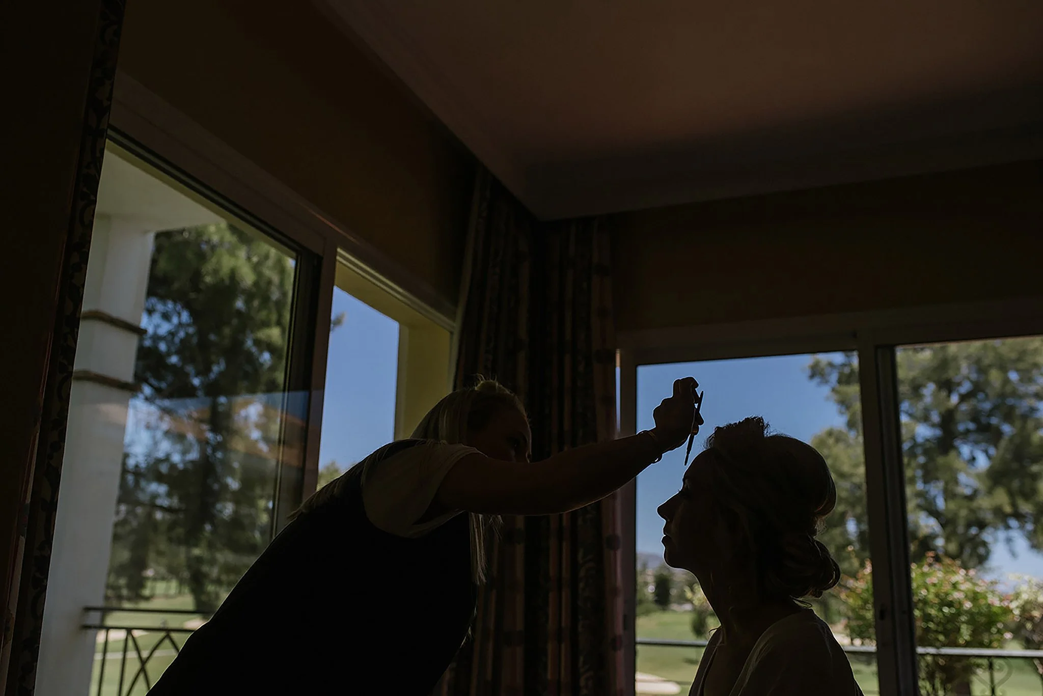 A person applying makeup or face paint to another woman’s face indoors, with large windows showing trees and a blue sky outside.