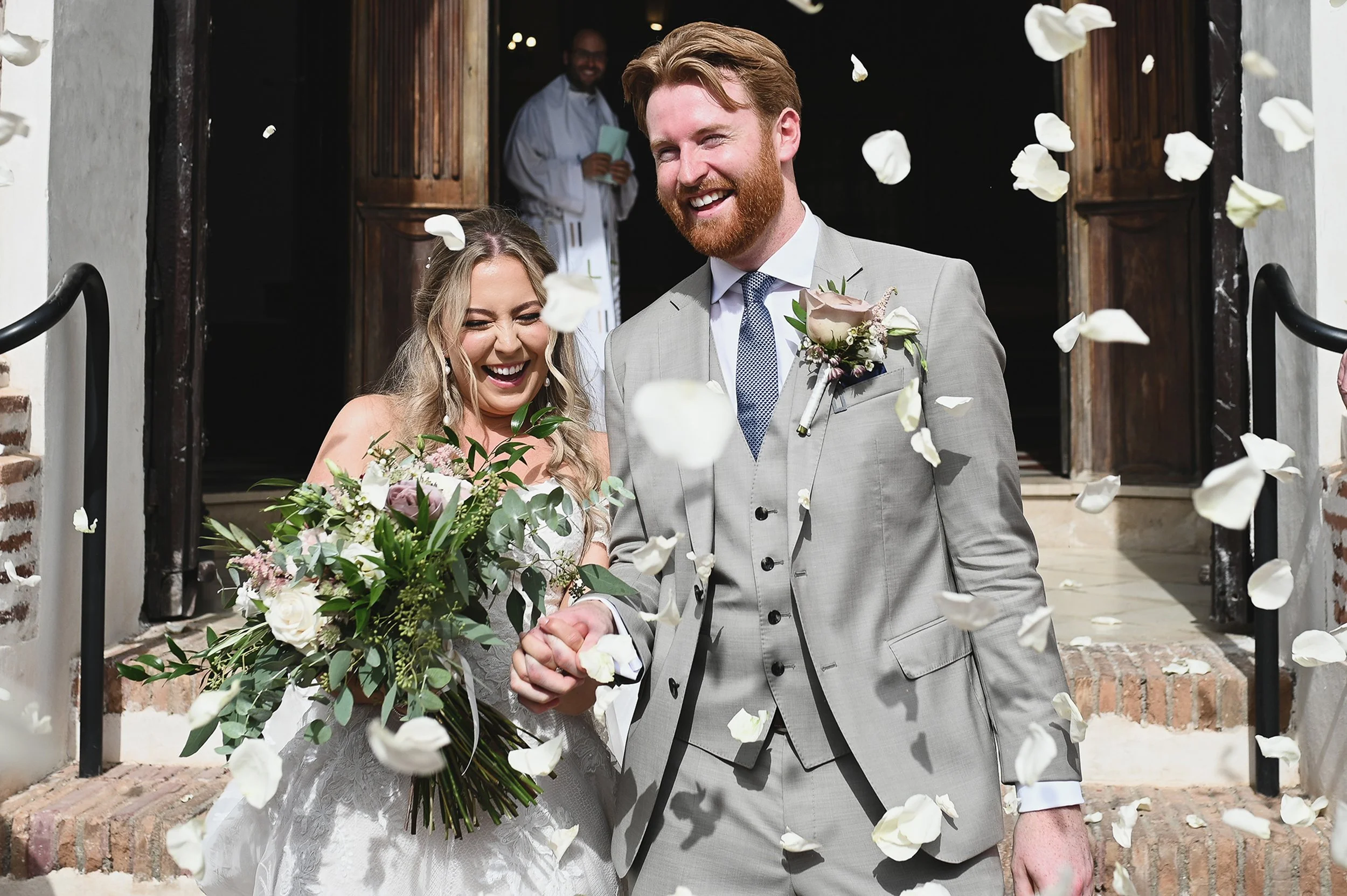 A newlywed couple exiting a wedding venue, surrounded by falling white flower petals. The bride is holding a large bouquet and laughing, while the groom is smiling and dressed in a light gray suit with a boutonniere. A man is seen in the background n