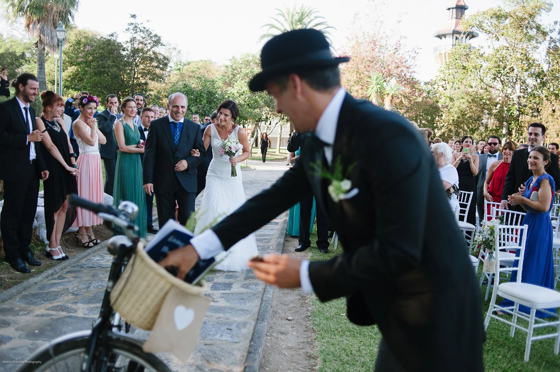 A wedding ceremony outdoors with a bride in a white dress walking down the aisle, surrounded by guests in colorful attire. A man in a black suit and hat is in the foreground, looking at his phone next to a bicycle with a basket. The background shows 