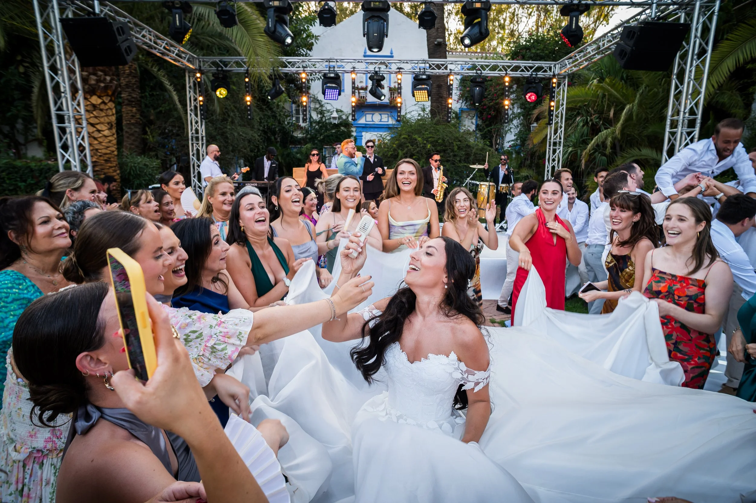 A wedding celebration with the bride in a white gown, surrounded by happy guests at an outdoor reception with a stage and band.