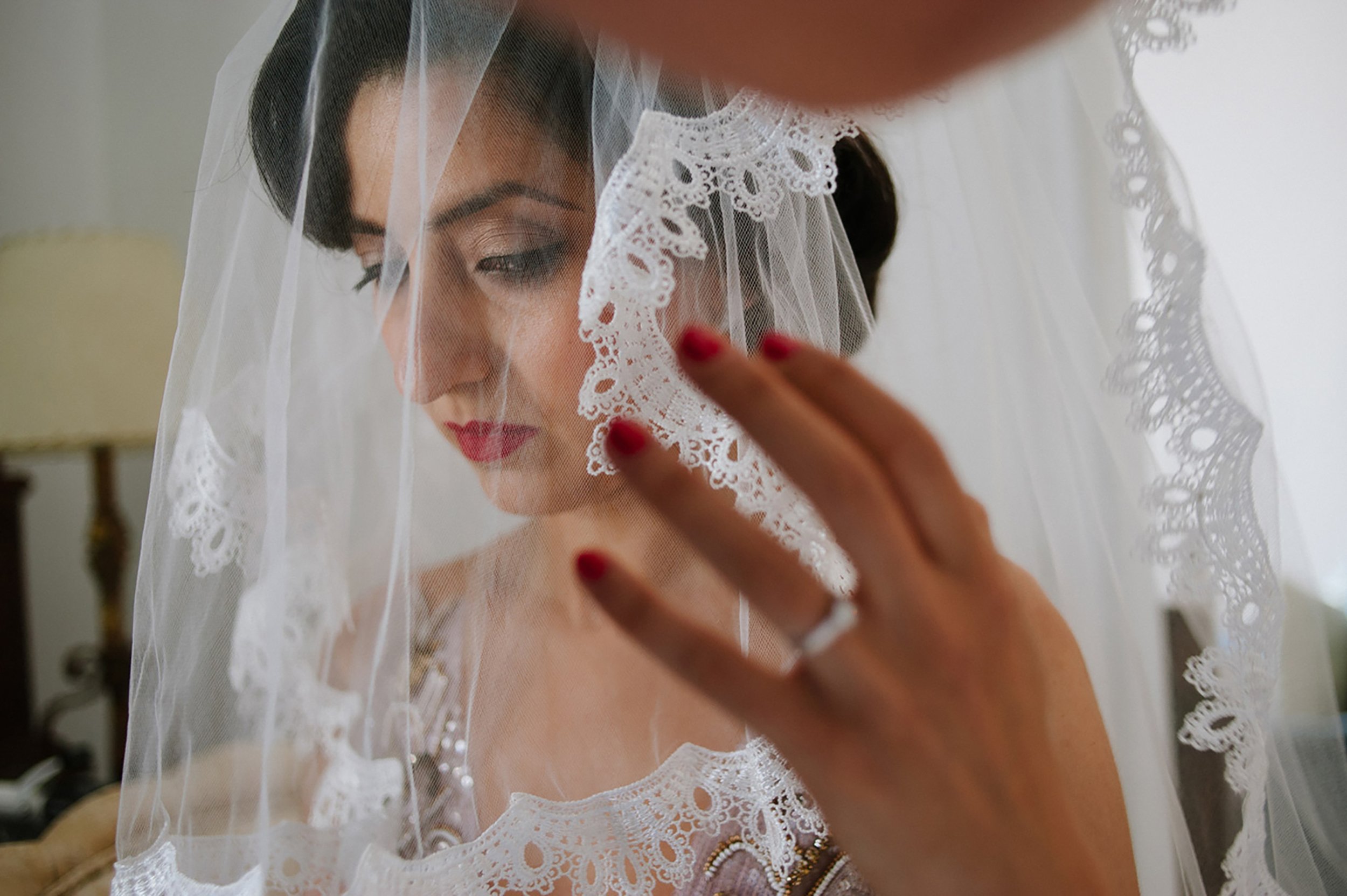 A woman wearing a lace veil and red lipstick, looking down as she adjusts her veil with her hand.