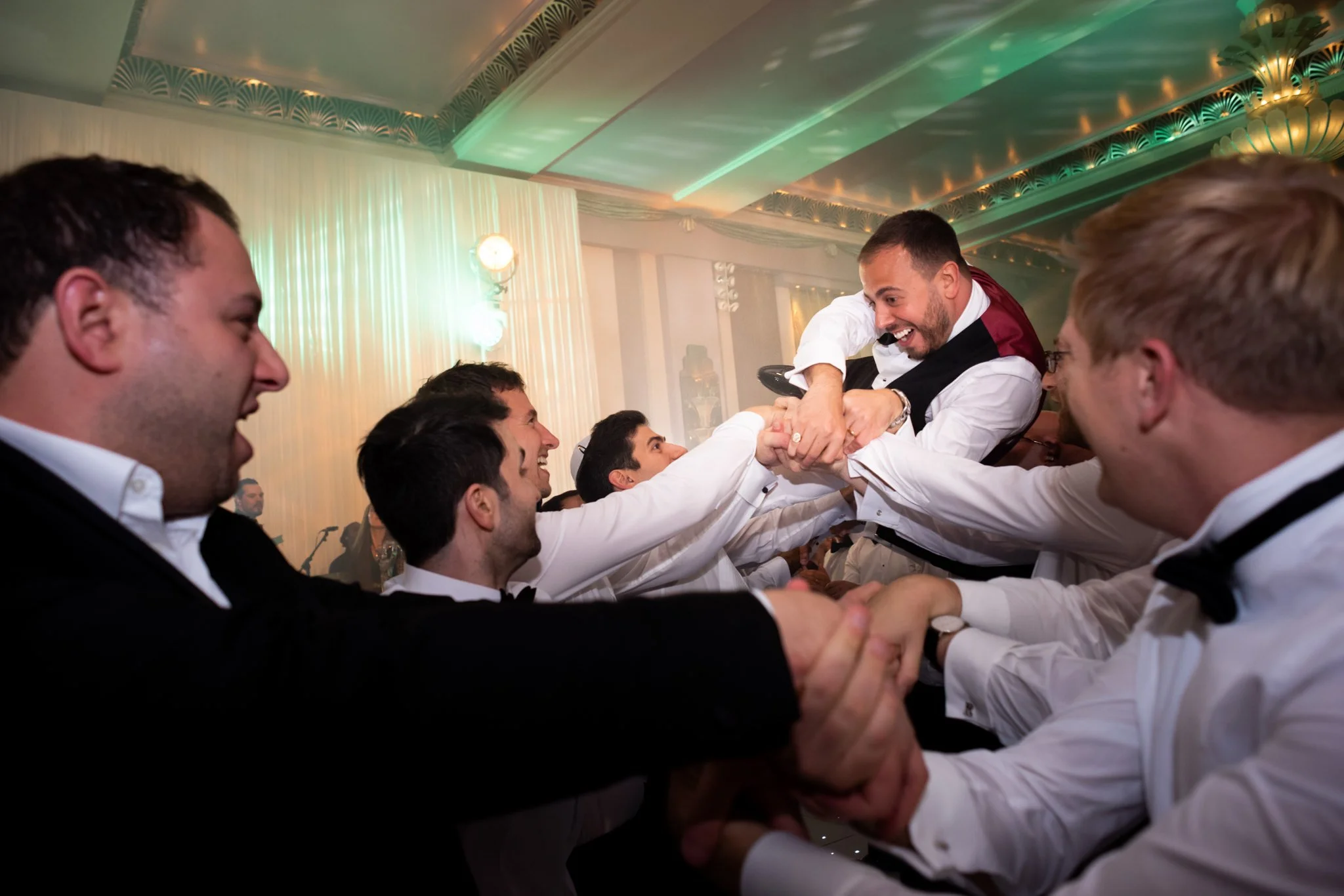 A groom being lifted and celebrated by groomsmen at a wedding reception, all smiling and laughing.