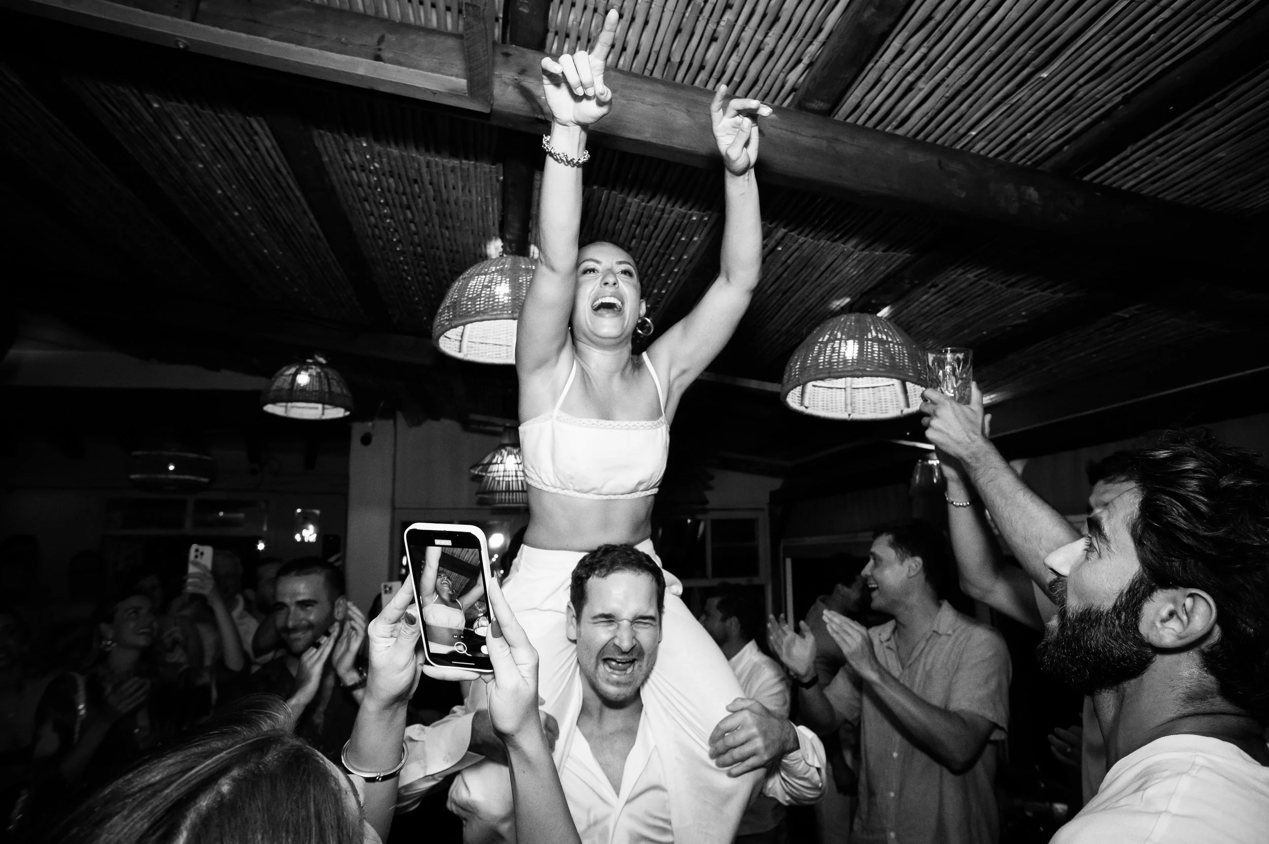 A celebration scene with a woman riding on a man's shoulders, raising her arms and smiling. Several people around are clapping, smiling, and some are taking photos at an indoor party with hanging lamps.