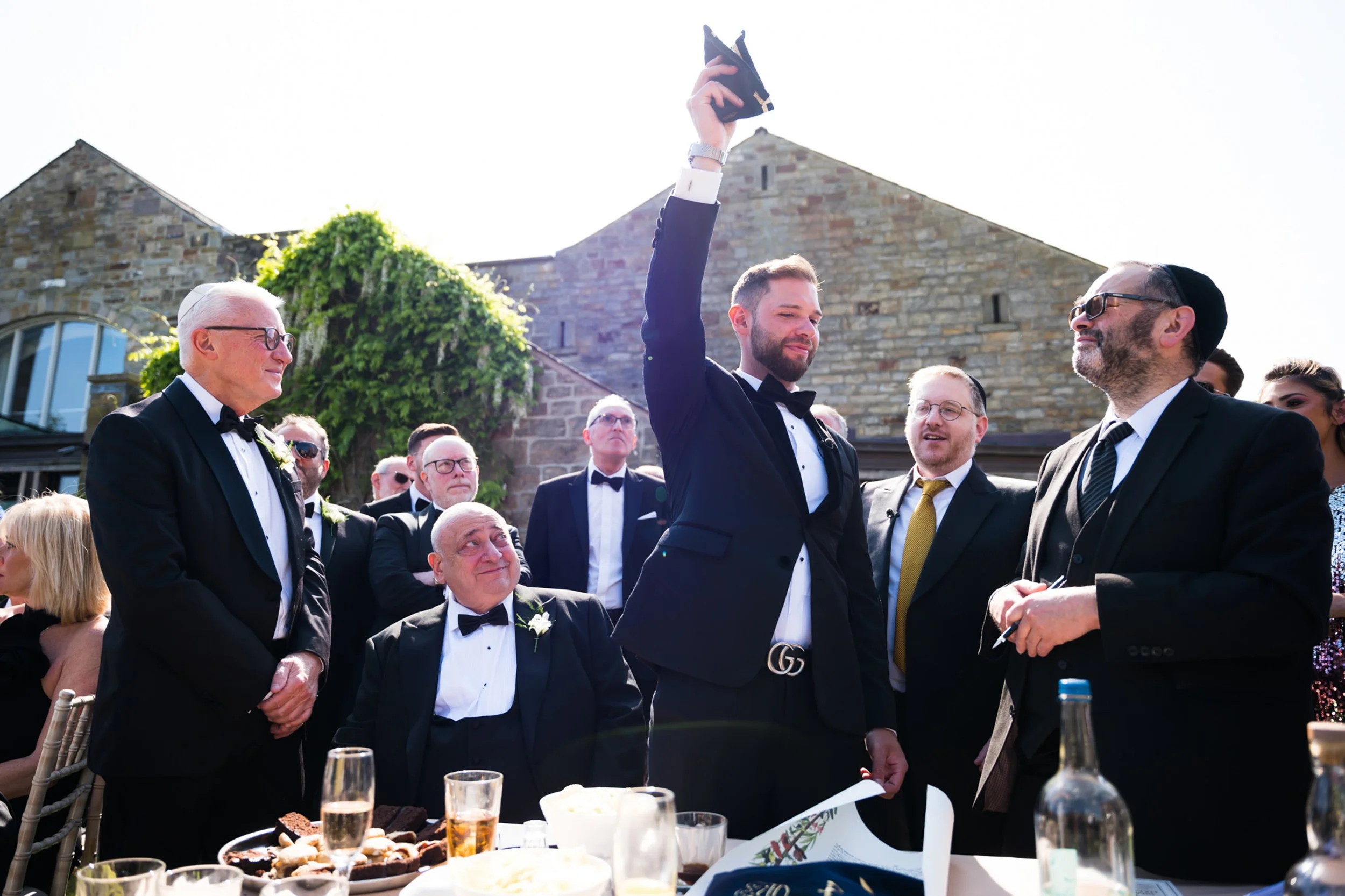 A man in a black tuxedo and bow tie raising his arm in celebration during a wedding reception outdoors, surrounded by guests also dressed in formal attire, with a stone building and vine-covered wall in the background.