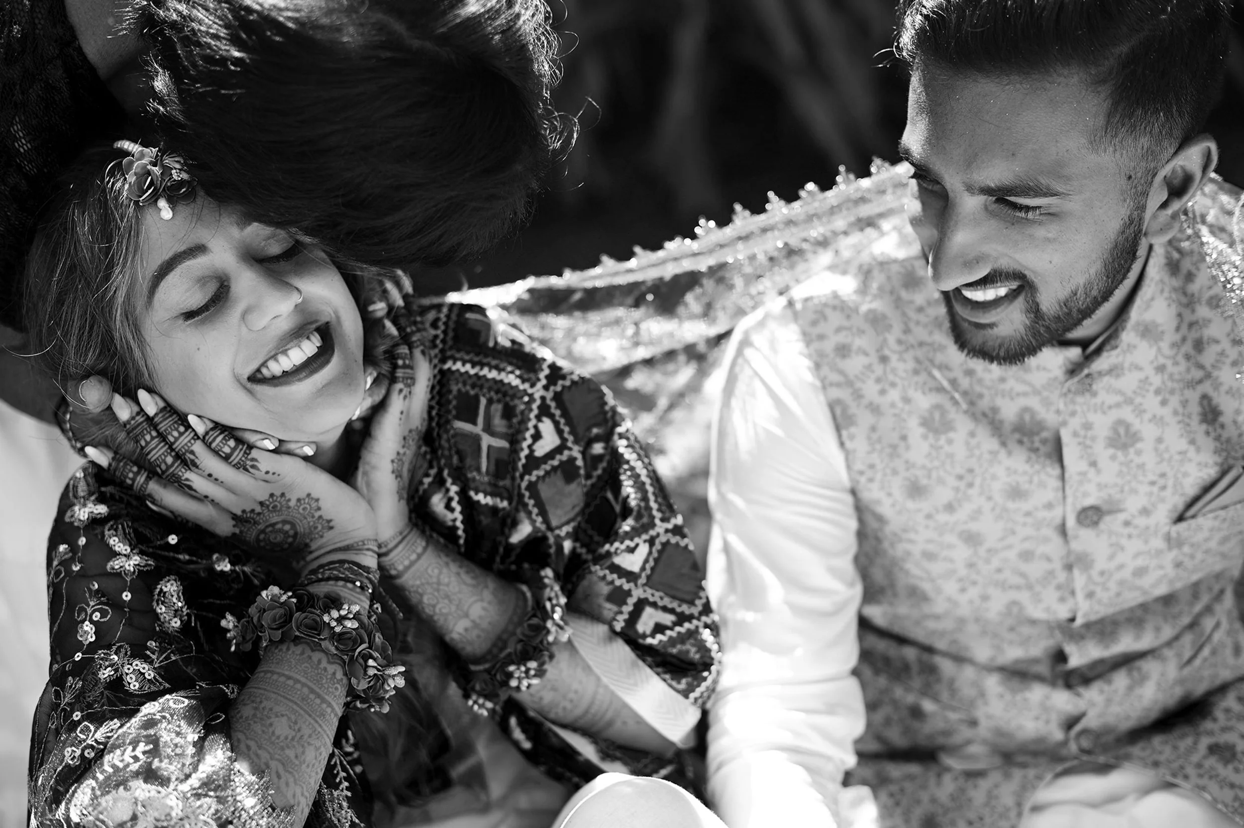 A woman and a man appear to be sharing a joyful moment, with the woman smiling and resting her head on the man's shoulder. The woman has traditional henna designs on her hands and is wearing jewelry and a patterned sari, indicating a celebratory or c