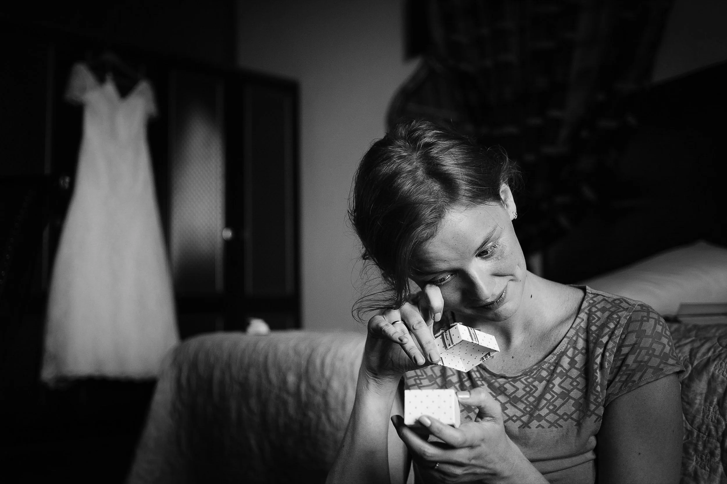 A woman with short hair opening a small gift box with a surprised expression in a dark room, with a wedding dress hanging in the background.