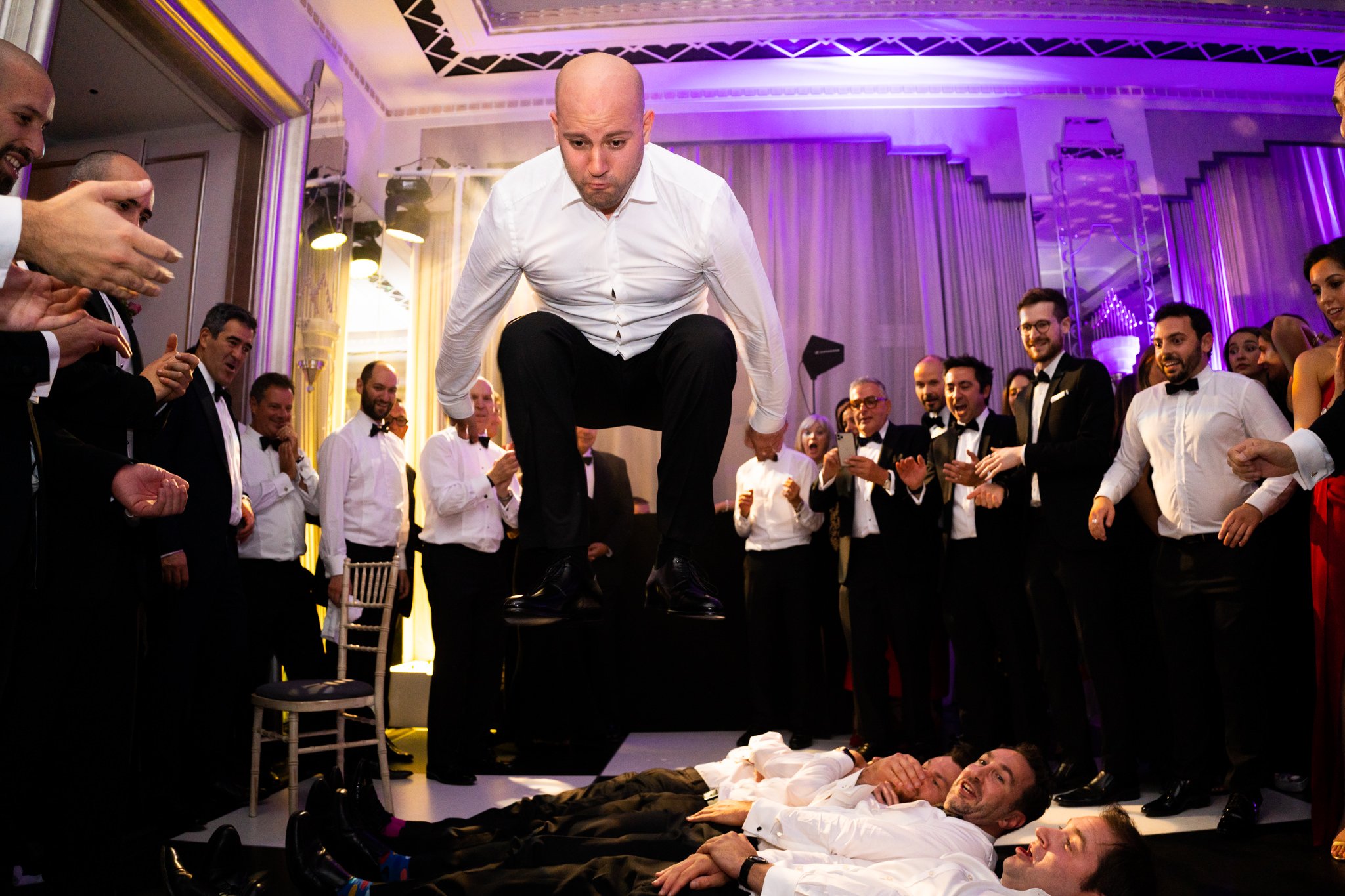 Guests at a formal event watch a man jump over other men lying on the floor, with many people smiling and taking photos.