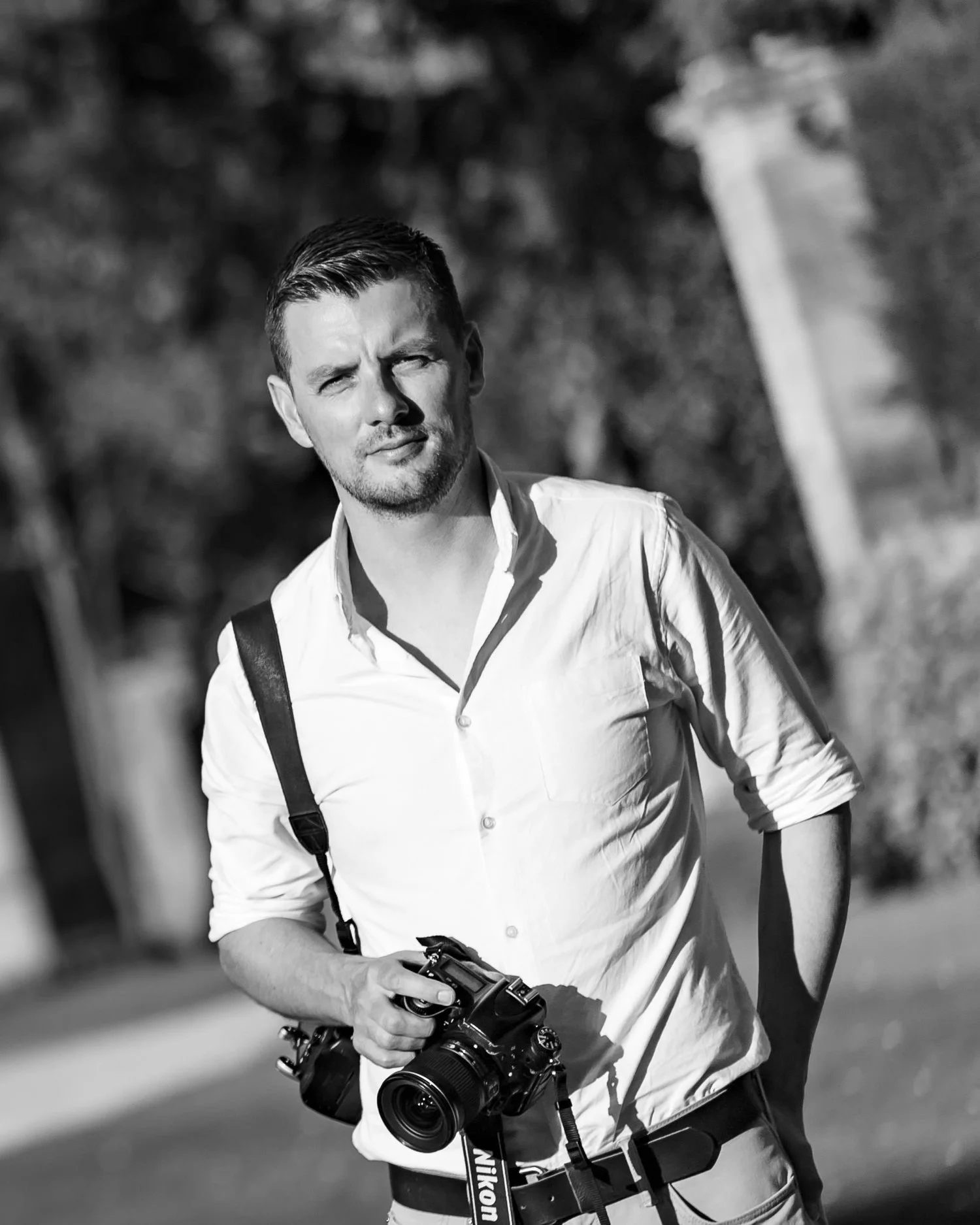 Black and white photo of a man with short hair and facial hair, wearing a light-colored button-down shirt with rolled-up sleeves, holding a Nikon camera with a strap, standing outdoors with trees and a pathway in the background.