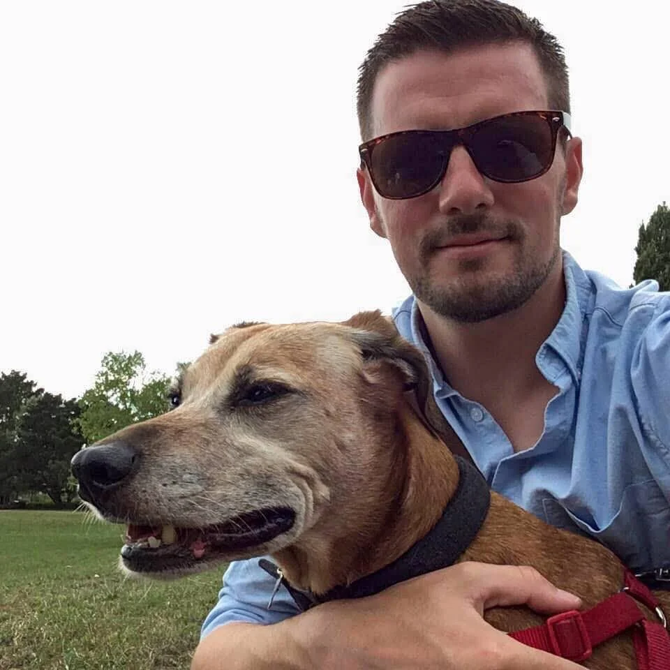 A man wearing sunglasses and a blue shirt taking a selfie with his dog outdoors in a park.