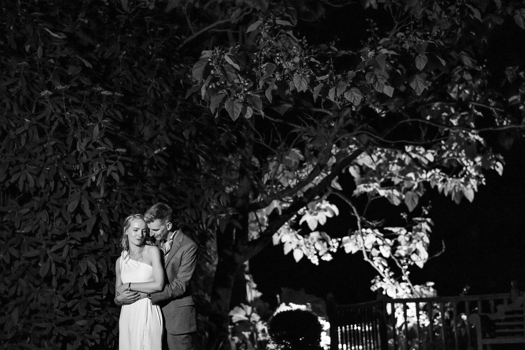 A black and white photo of a couple embracing under a large tree at night, with a fence in the background.