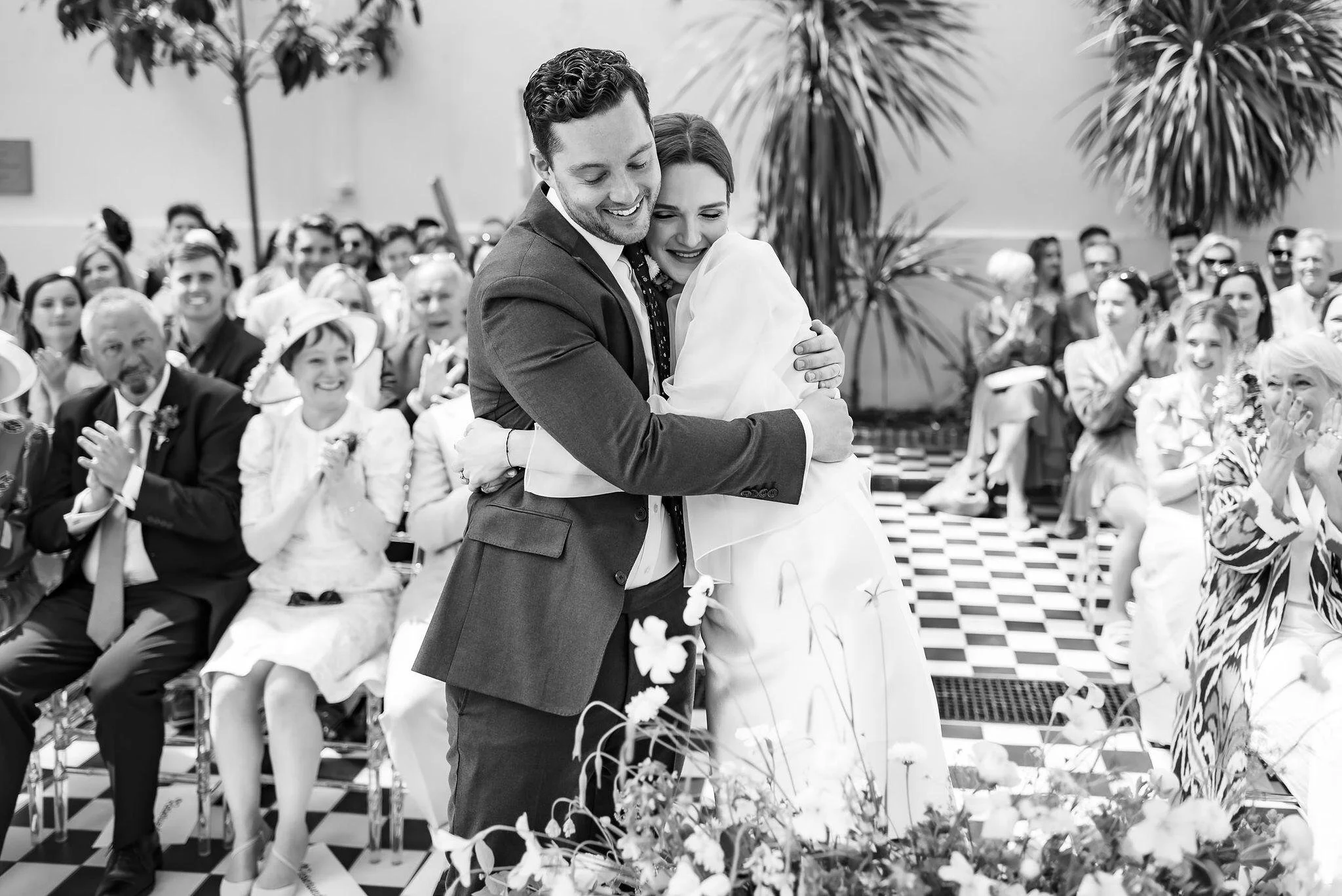 A black and white photo of a man and woman hugging in the center during a wedding ceremony, surrounded by seated and standing guests clapping and smiling.