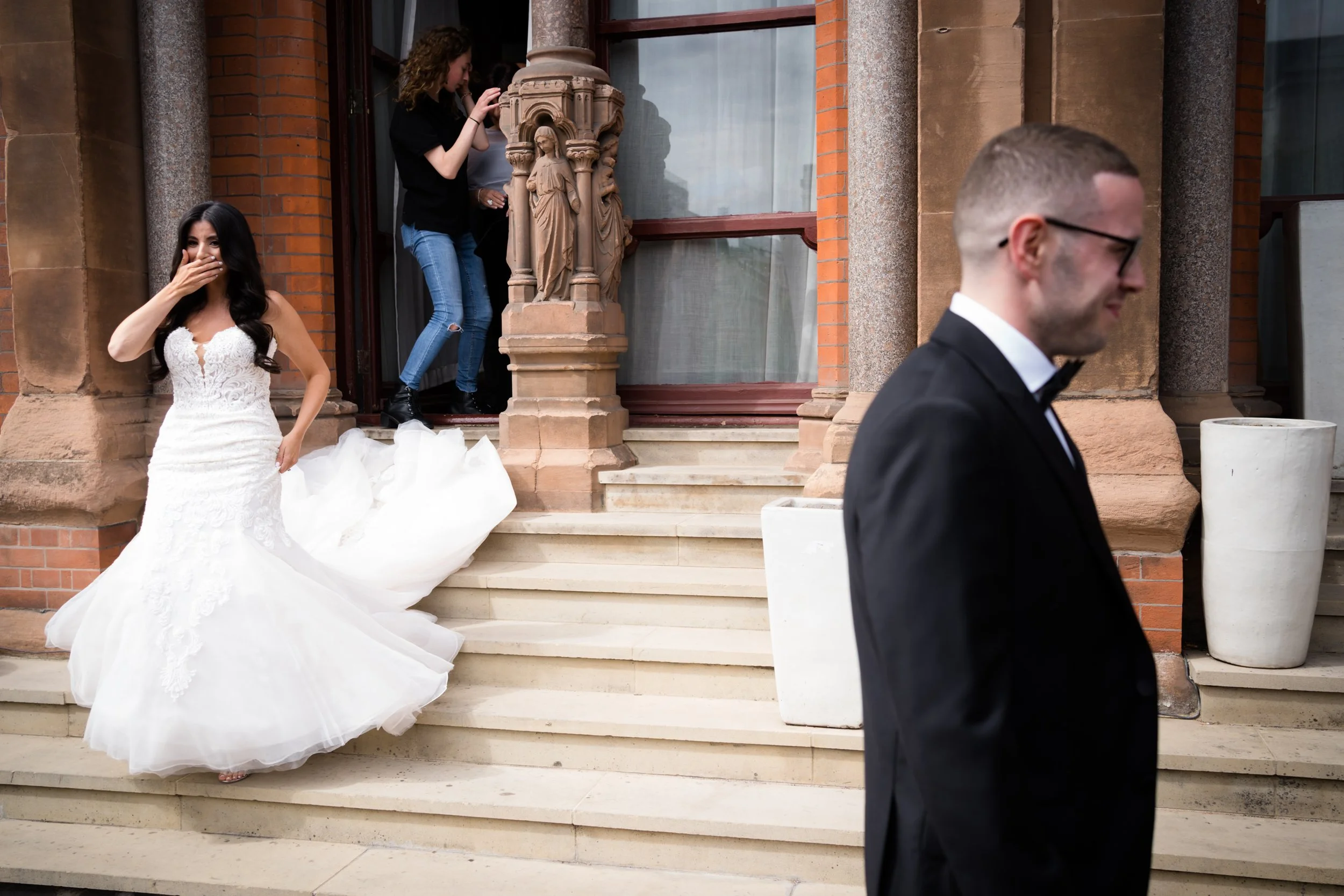 A bride in a white wedding gown with lace details stands on steps outside a brick building, covering her mouth with her hand and smiling, while a man in a tuxedo walks past with his face out of focus. Two women, one taking a photo or video, are at th