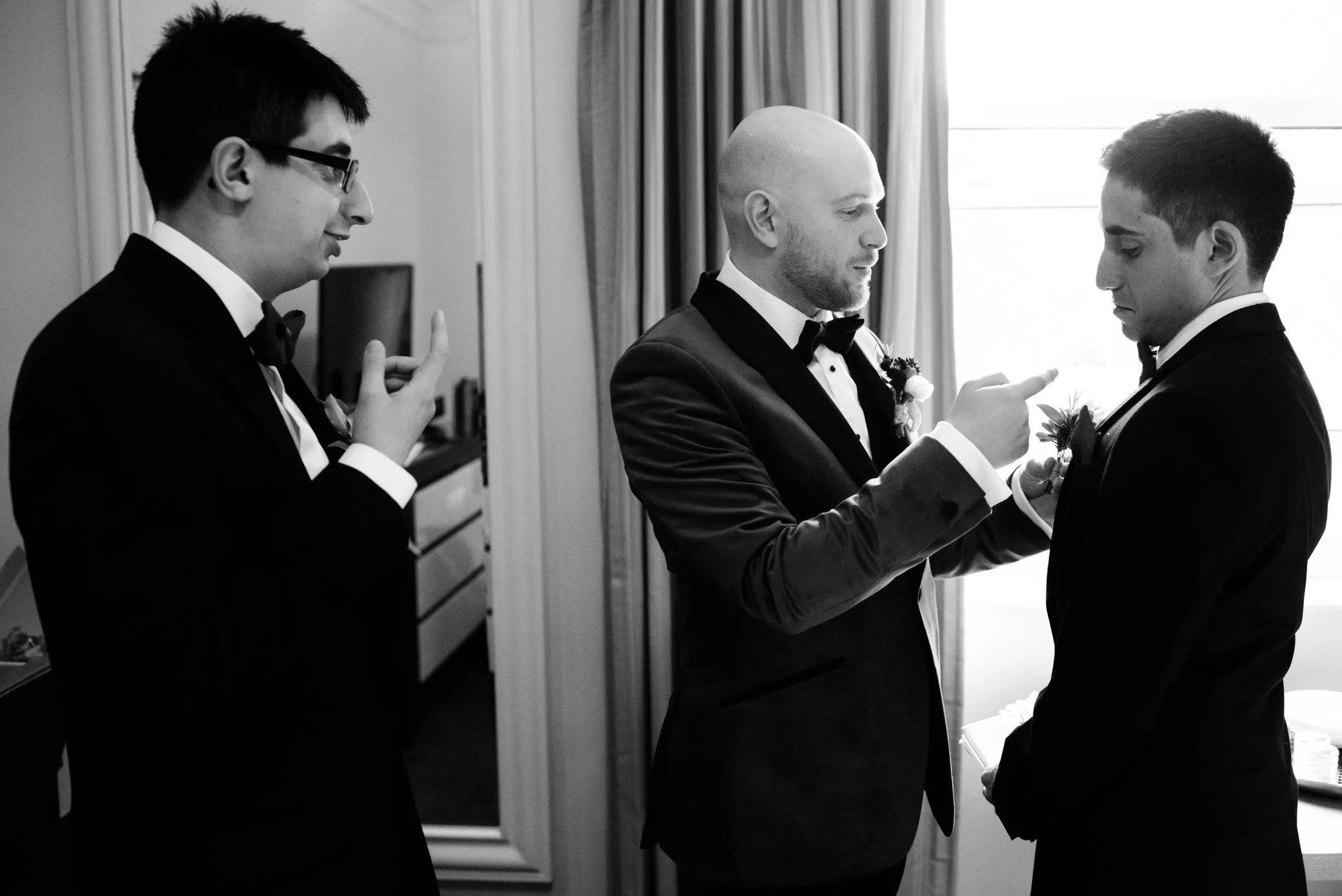 Three men in black tuxedos and bow ties, two with boutonnières, in a room with curtains and a mirror, one bald man pointing at the chest of a younger man, possibly during a wedding preparation.