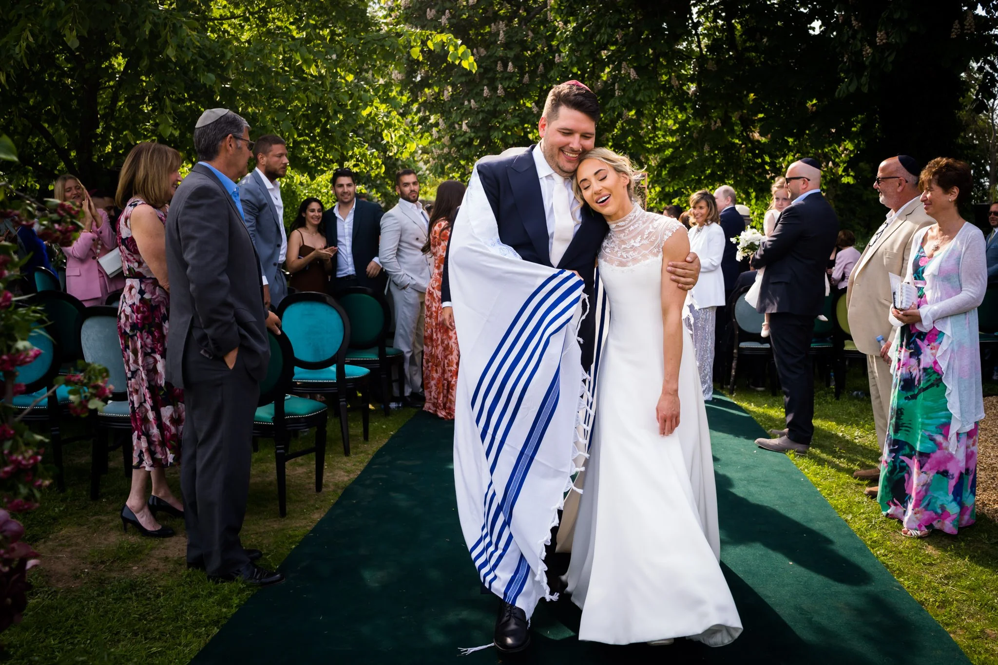 A newly married couple, the groom in a dark suit and the bride in a white wedding dress, are embracing and smiling during their outdoor wedding ceremony surrounded by guests.