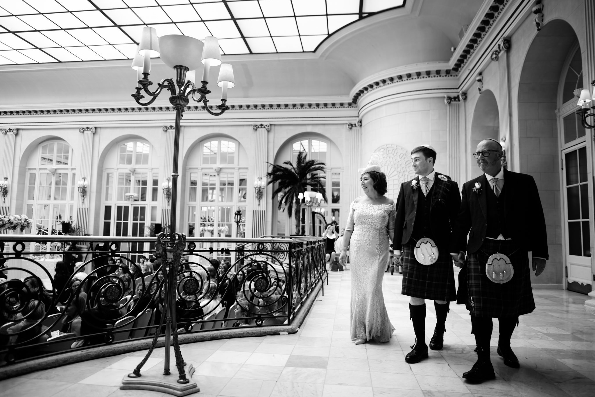 A bride and two men dressed in traditional Scottish kilts walking arm-in-arm inside an elegant hall with high ceiling, large windows, ornate chandeliers, and decorative railings during a wedding ceremony.