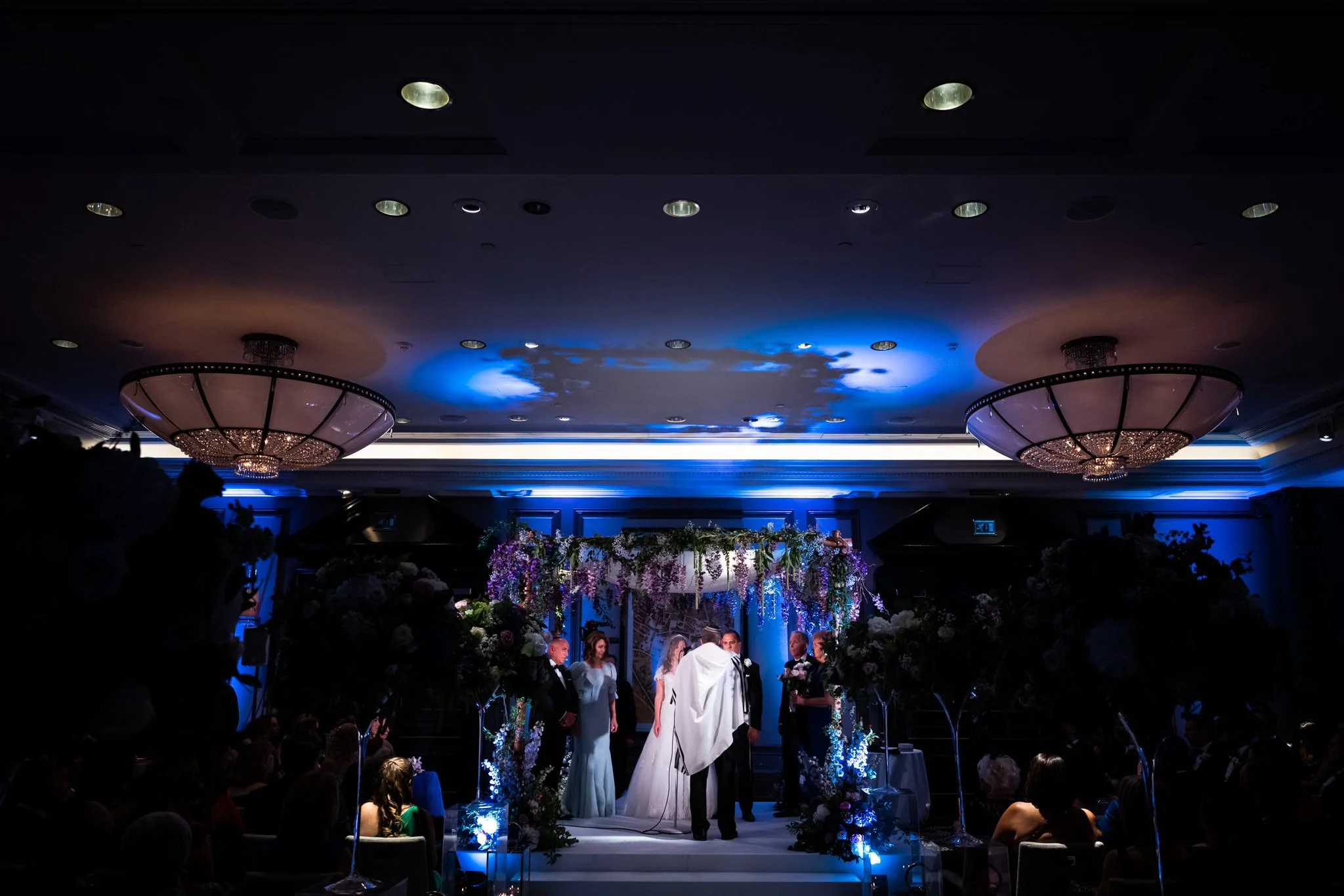 A wedding ceremony with the bride and groom standing under an arch decorated with flowers, in a dimly lit room with blue lighting and chandelier ceiling fixtures.