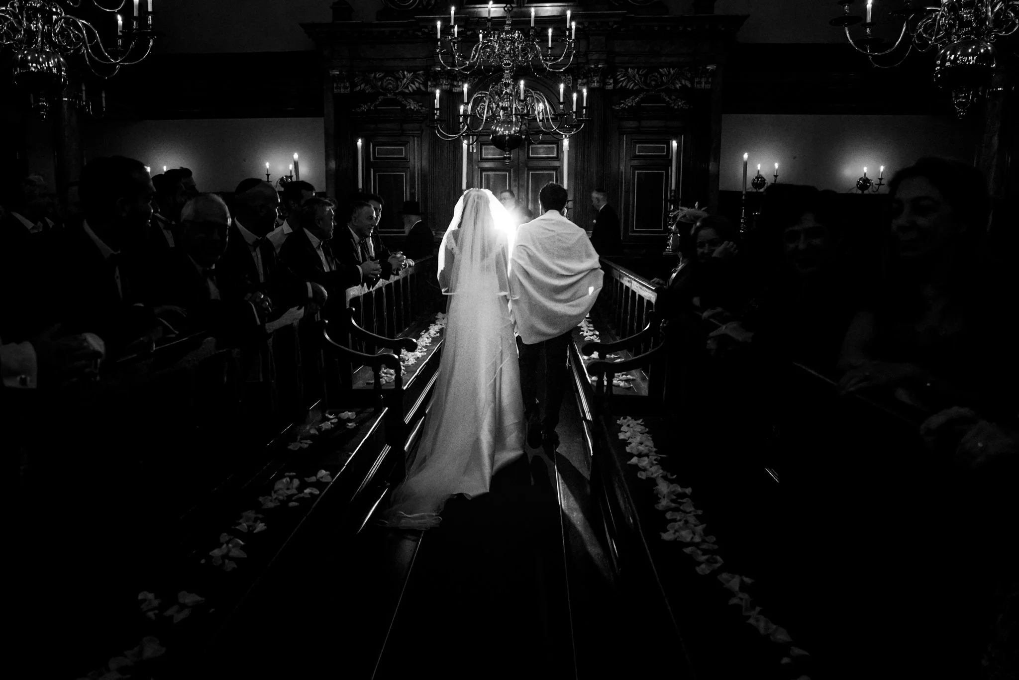 A black and white photo of a wedding ceremony in a church, with a bride and groom walking down the aisle, surrounded by seated guests on both sides, with flower petals scattered along the aisle.