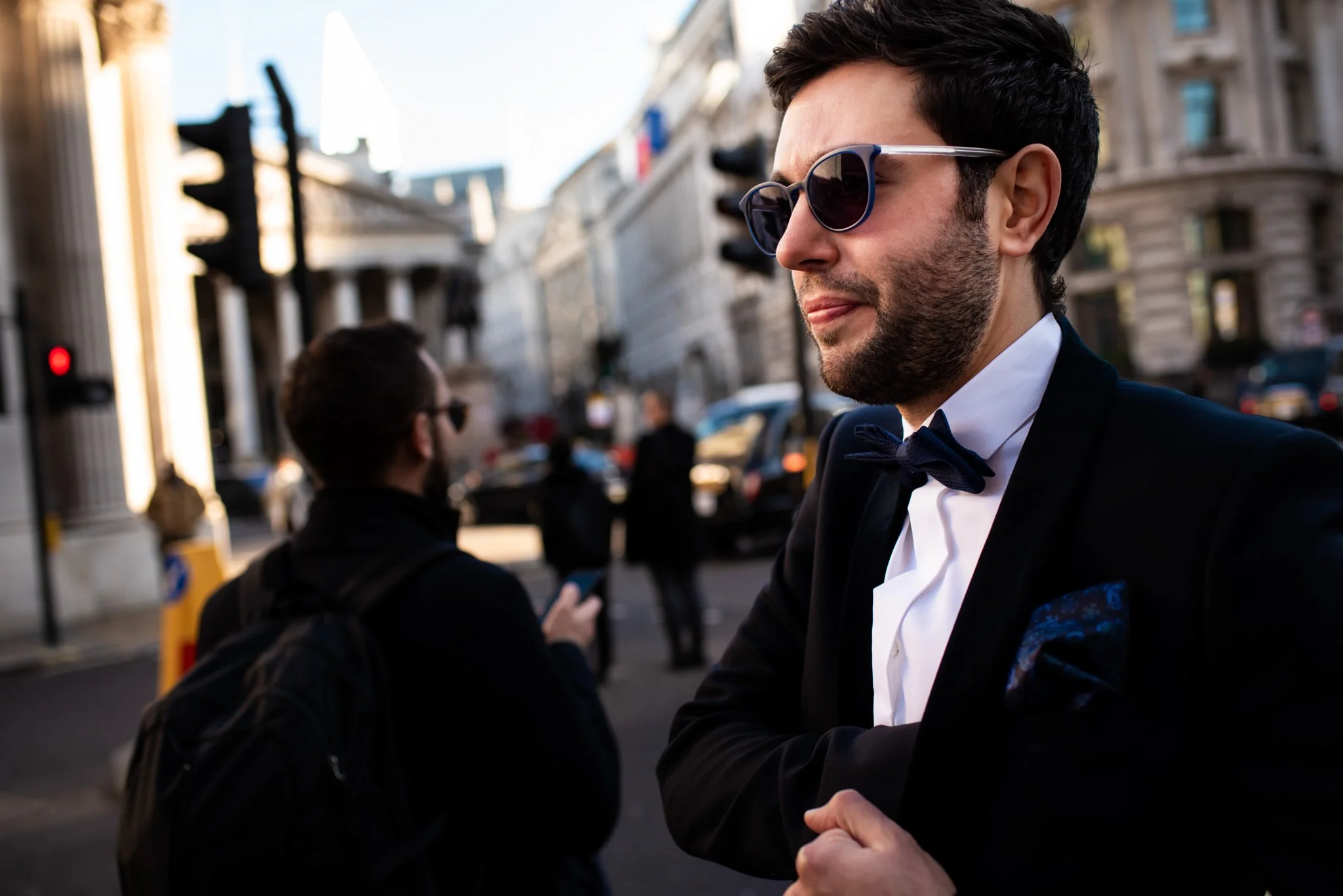 A man in a tuxedo with sunglasses standing on a city street during daylight.