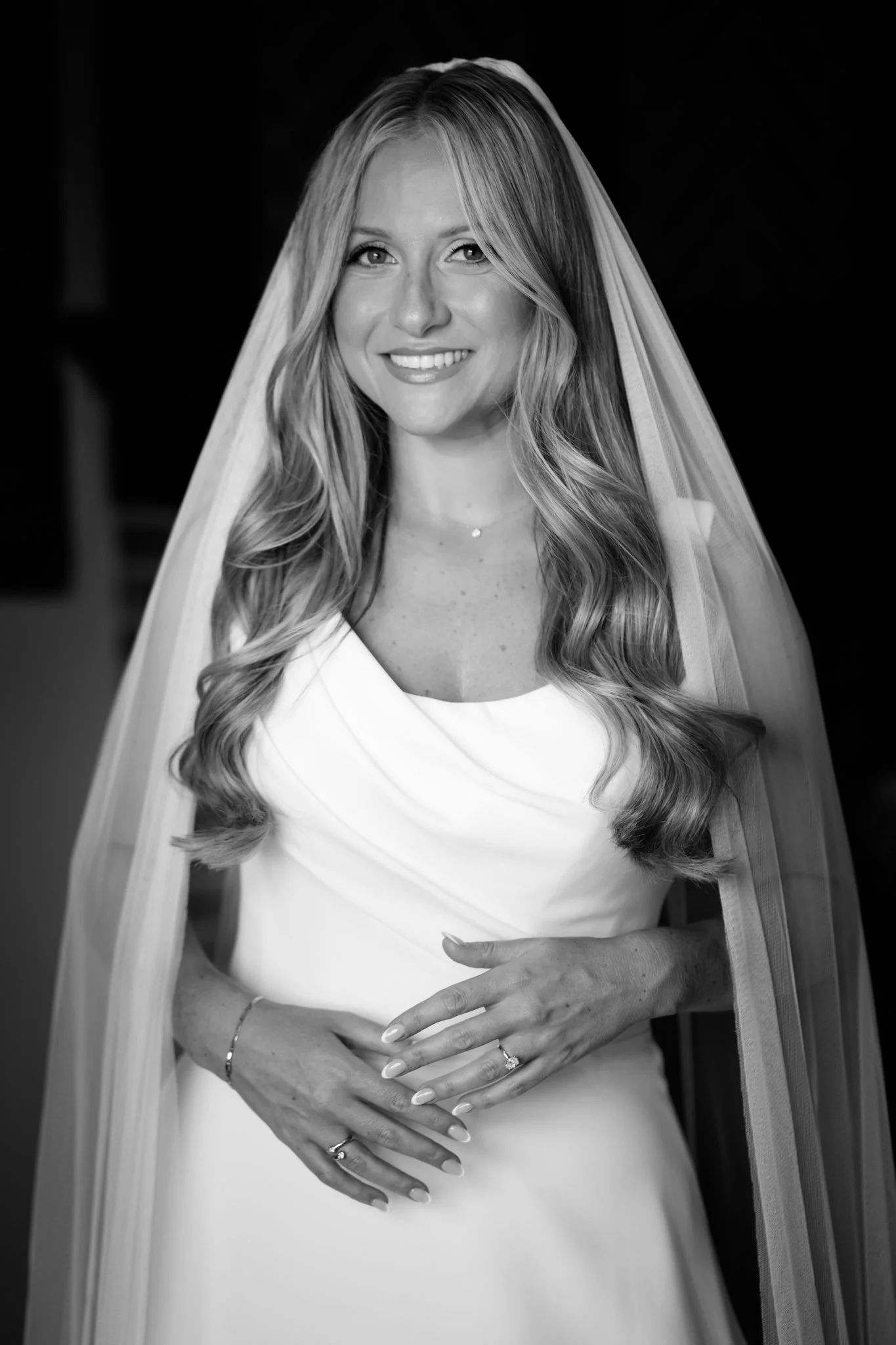 Black and white photo of a smiling woman in a wedding dress with a veil, holding her hands over her stomach, showing wedding rings.